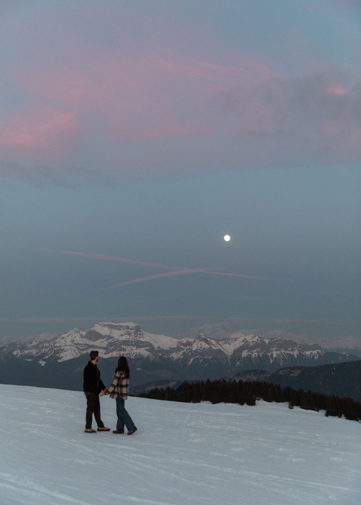 Silhouettes of skiers on a snowy slope at dusk with majestic snow-capped mountains and a full moon in the background.