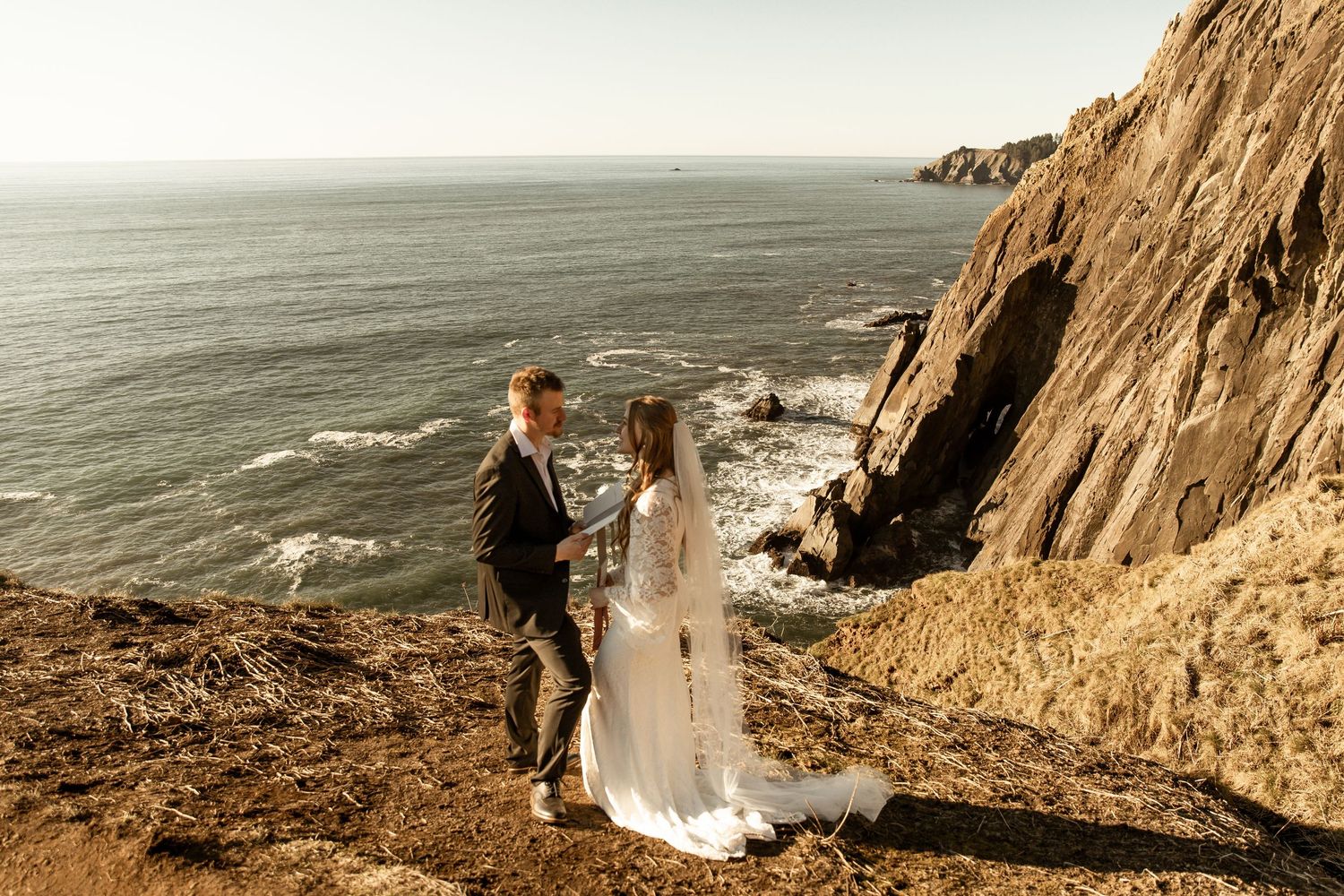 A couple stands at the edge of a dramatic coastal cliff overlooking the ocean during their wedding ceremony.