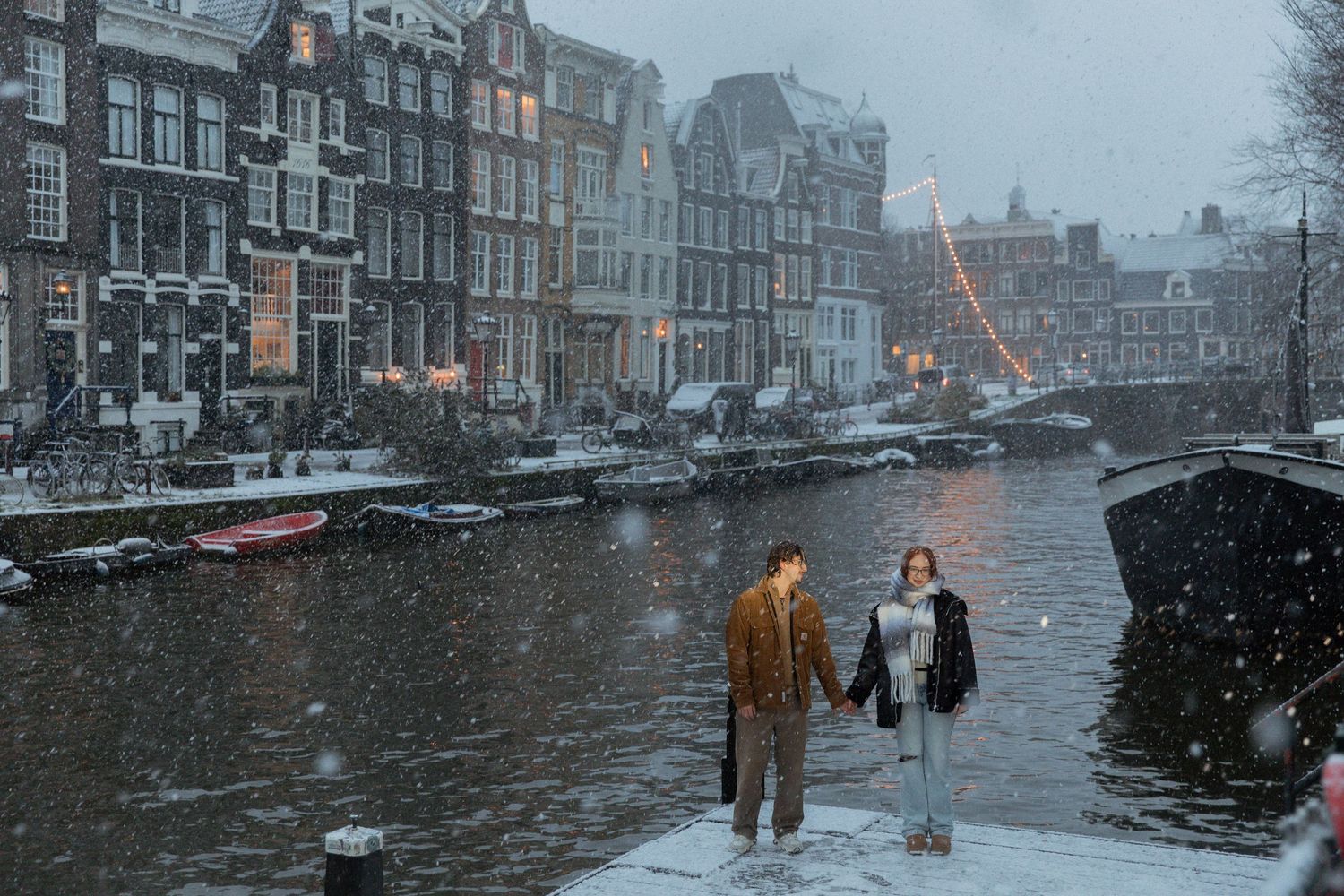 Snow falls over Amsterdam's iconic canal houses on a winter afternoon.