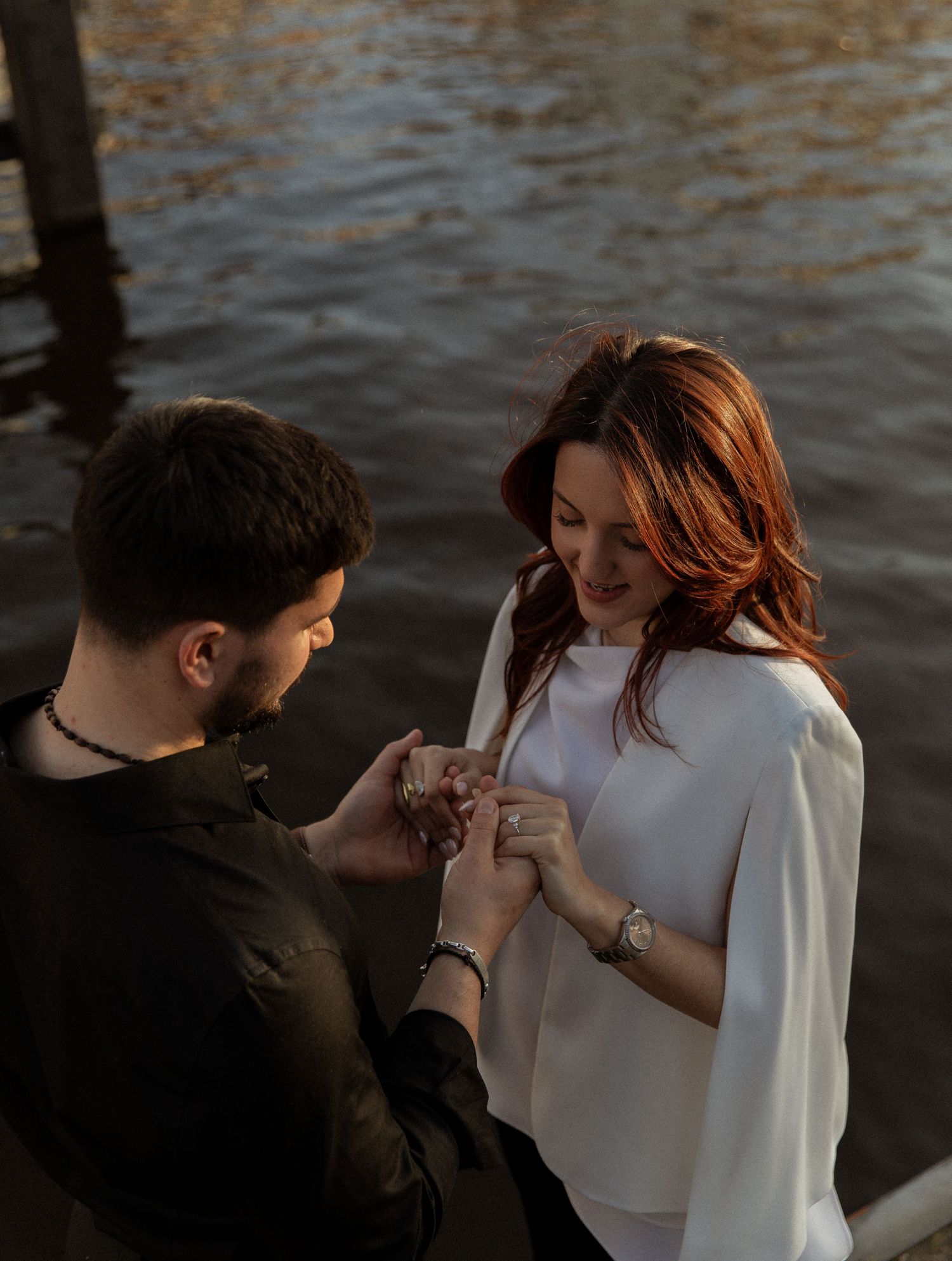Man kneeling to place engagement ring on womans finger by waterfront at golden hour.