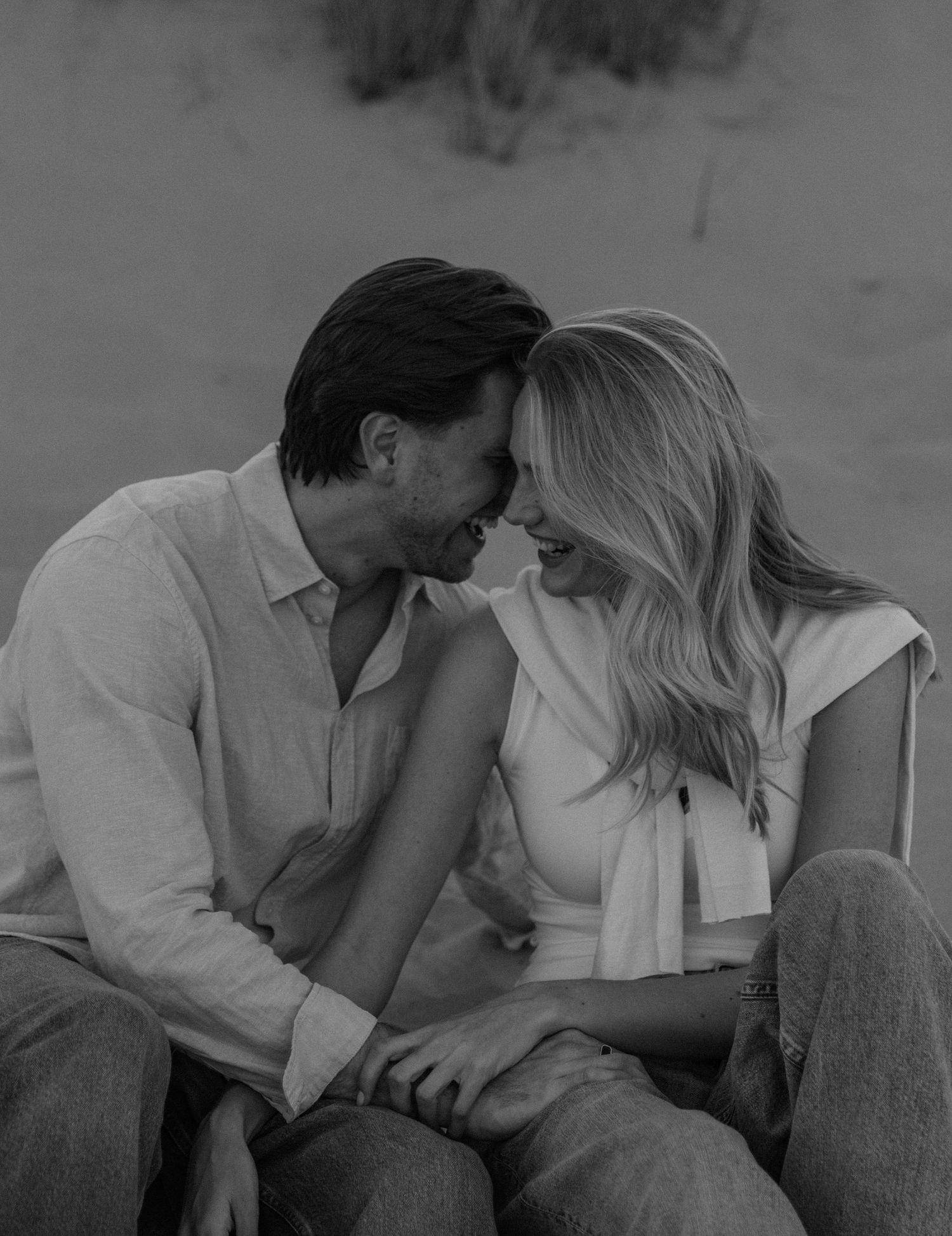 Black and white photo of a romantic couple sitting together on sand dunes, leaning in close and smiling.