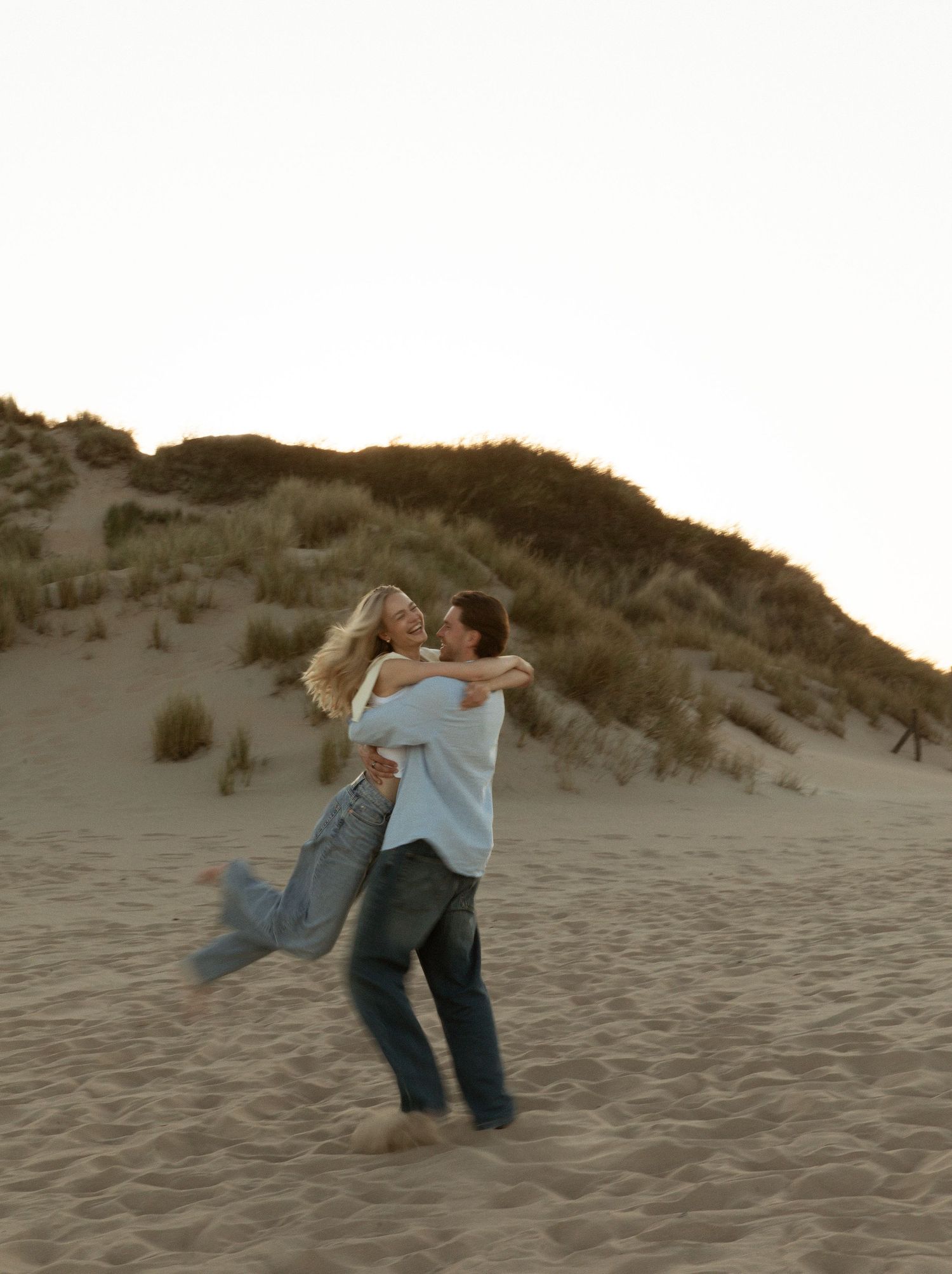A couple embracing and laughing on sandy dunes at sunset, with the man lifting the woman joyfully.