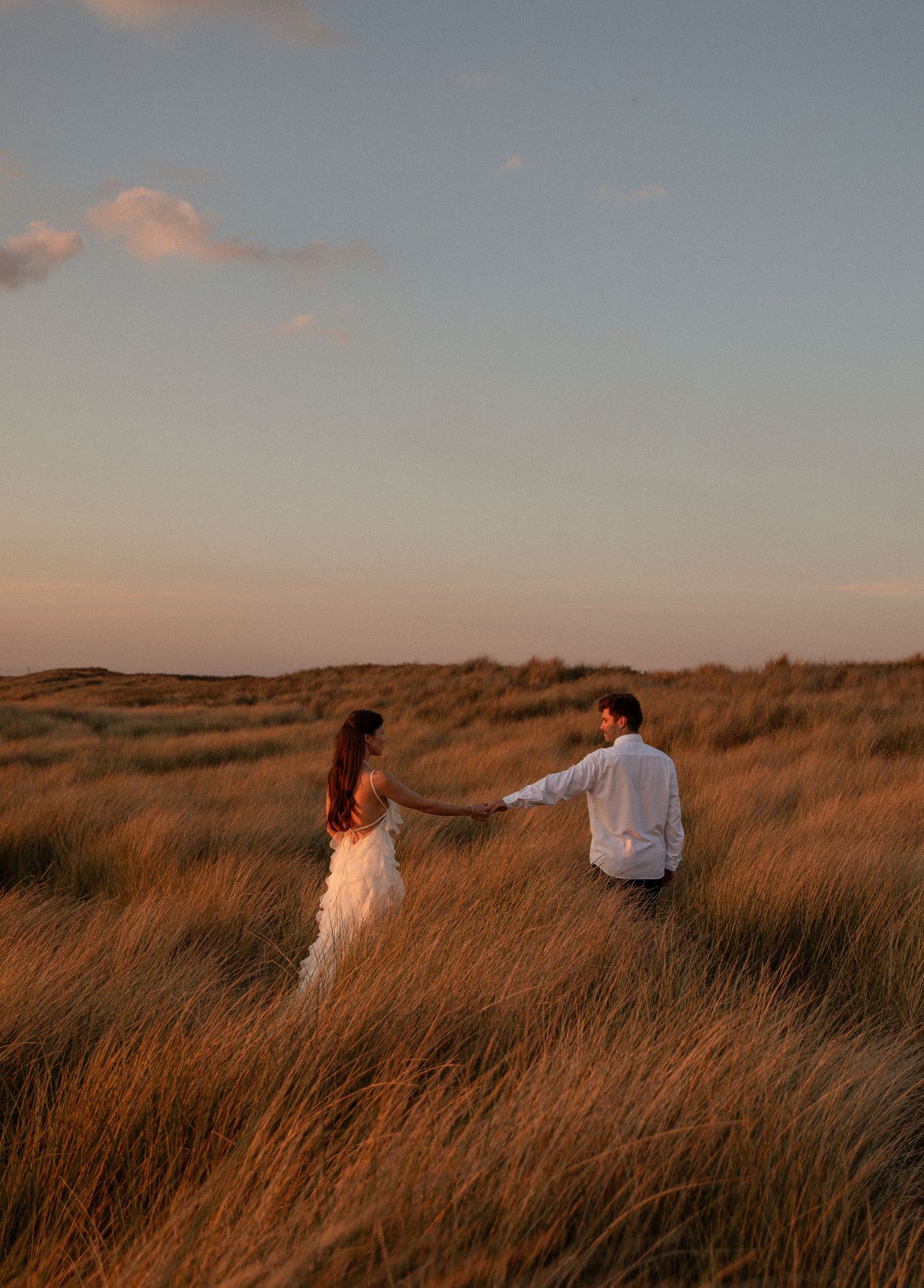Couple holding hands in golden grass fields at sunset, woman in white dress, romantic outdoor photography.