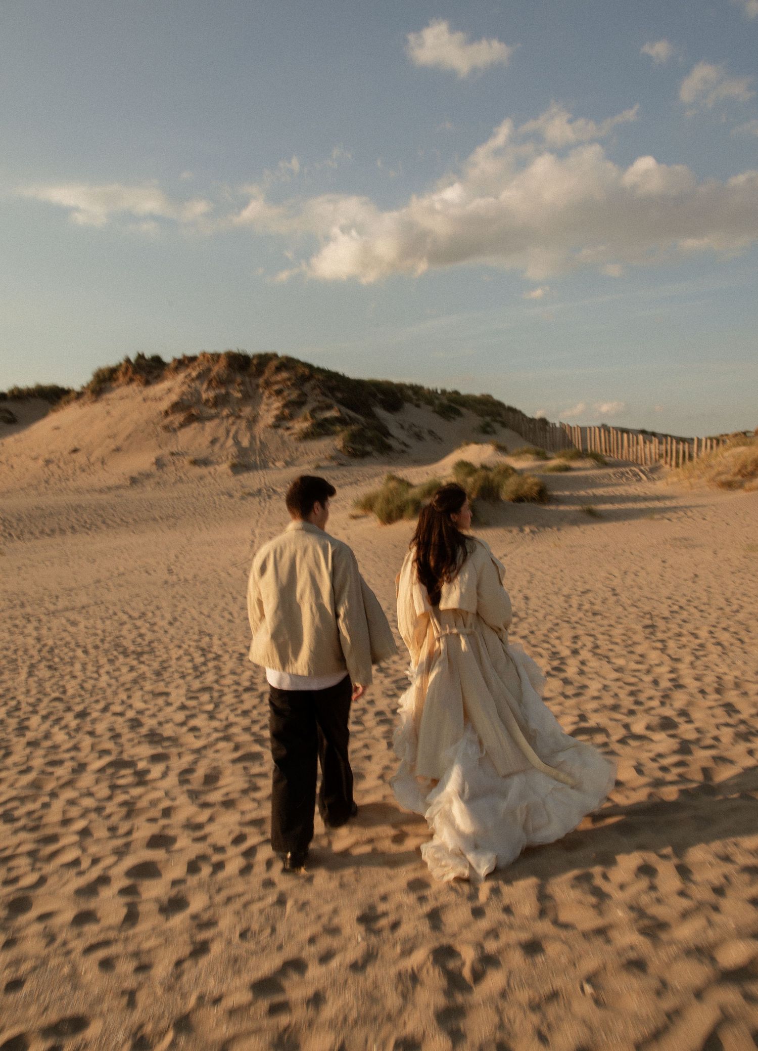 A couple in wedding attire walks hand-in-hand across a sandy beach toward golden dunes at sunset.