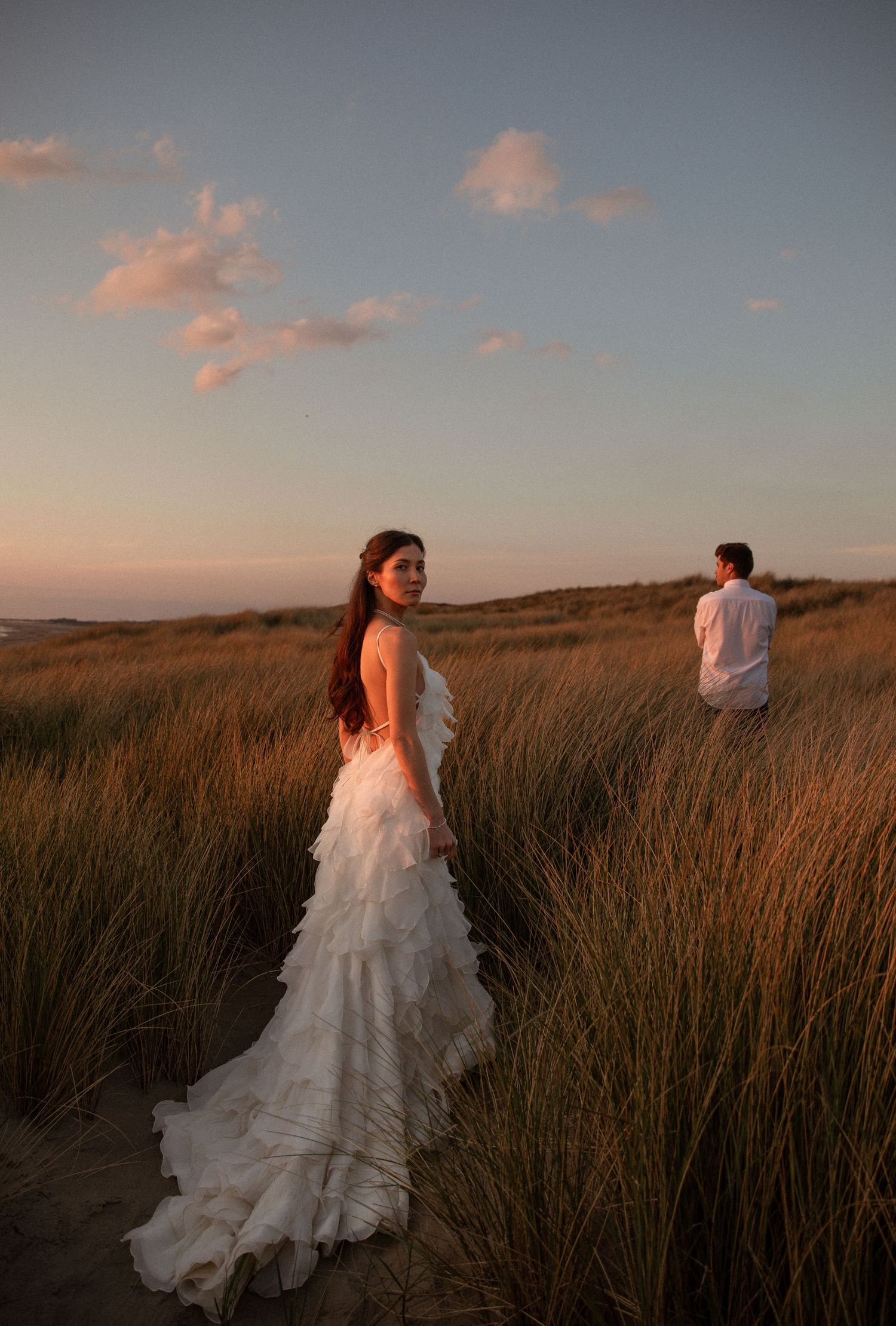 Bride in white ruffled gown stands in golden dune grass at sunset, groom in background during beach wedding shoot.