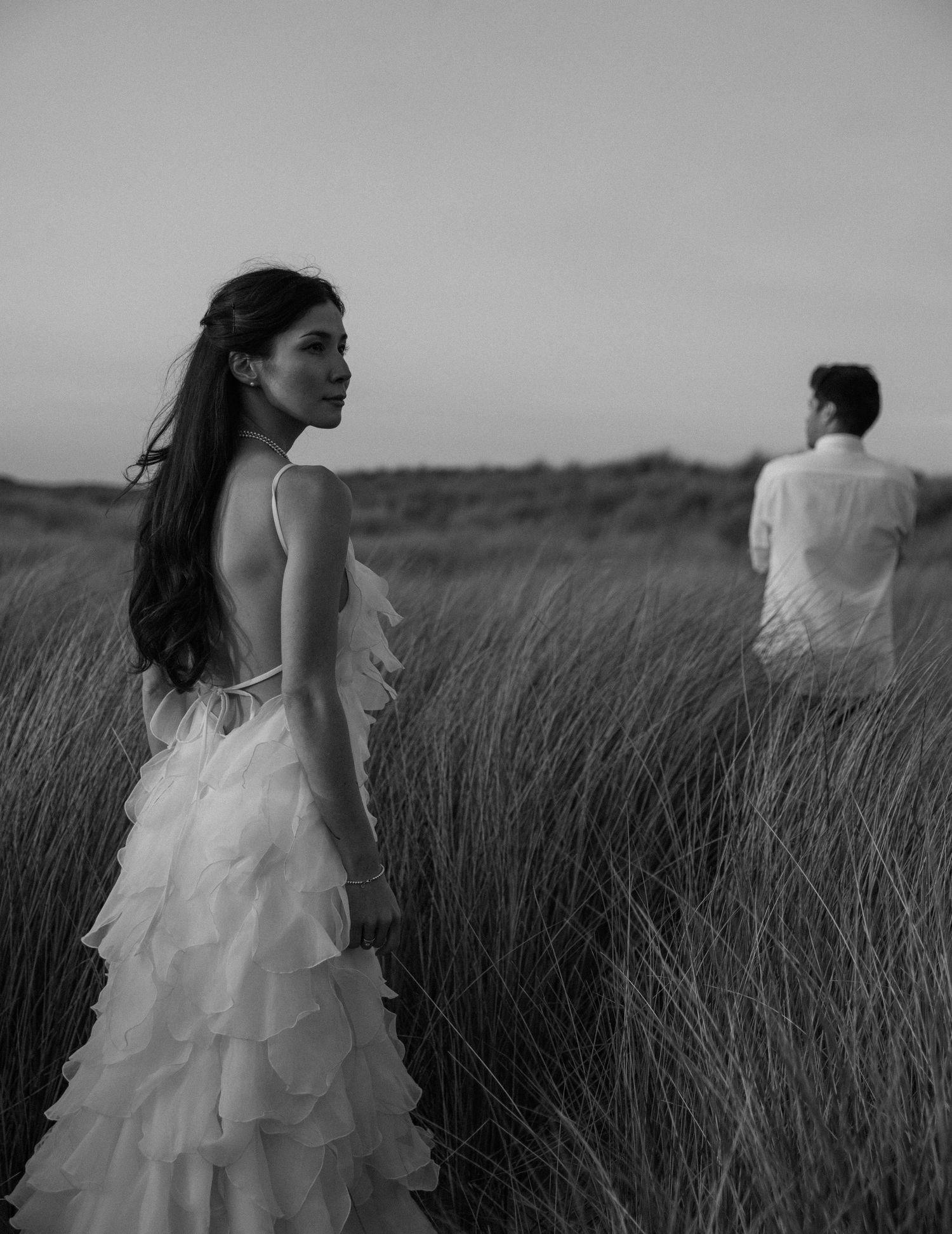 Black and white photo of a bride in a ruffled backless gown standing in a grass field with groom in background.