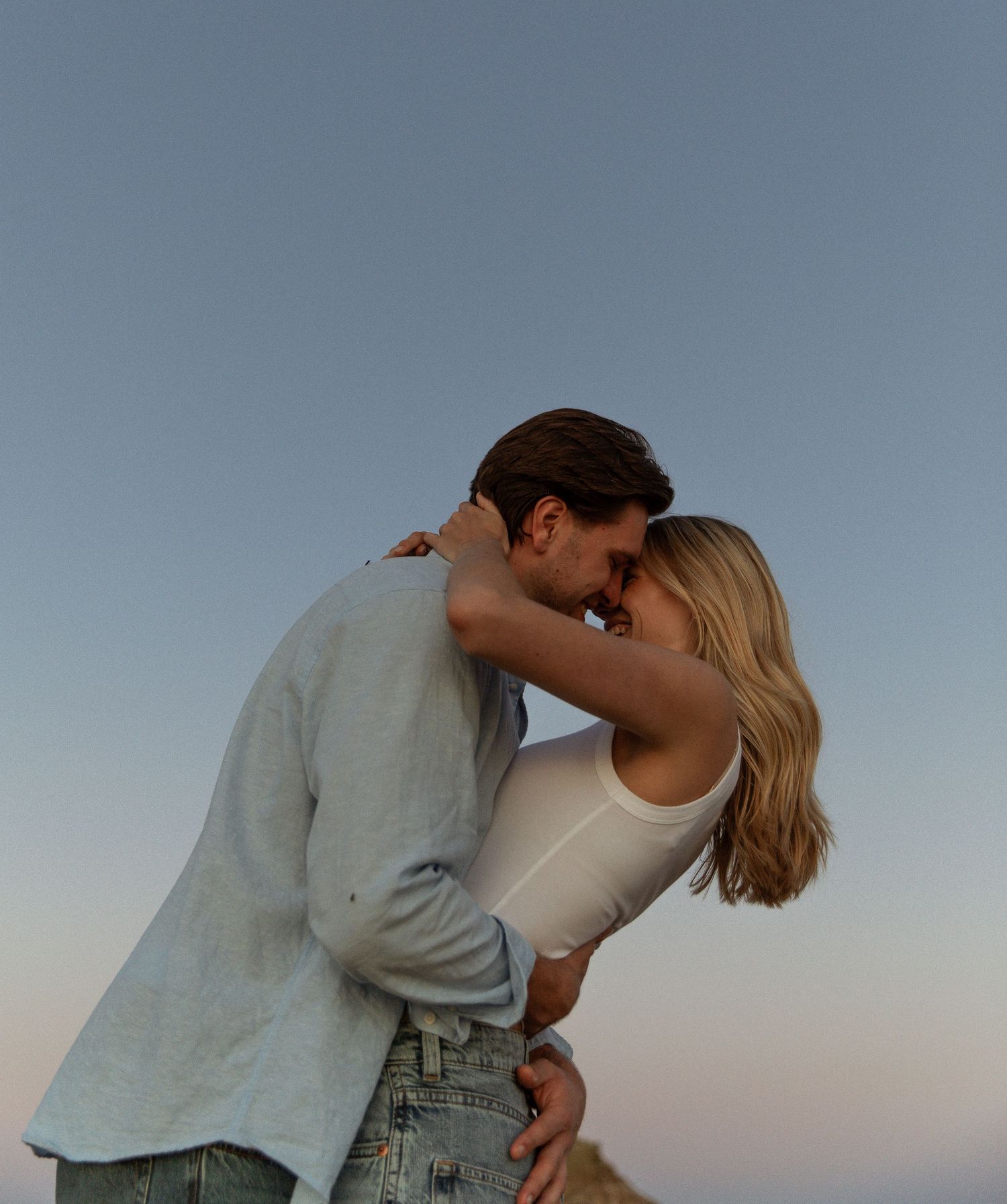 Romantic couple sharing a tender moment at sunset, embracing outdoors against a soft blue sky.