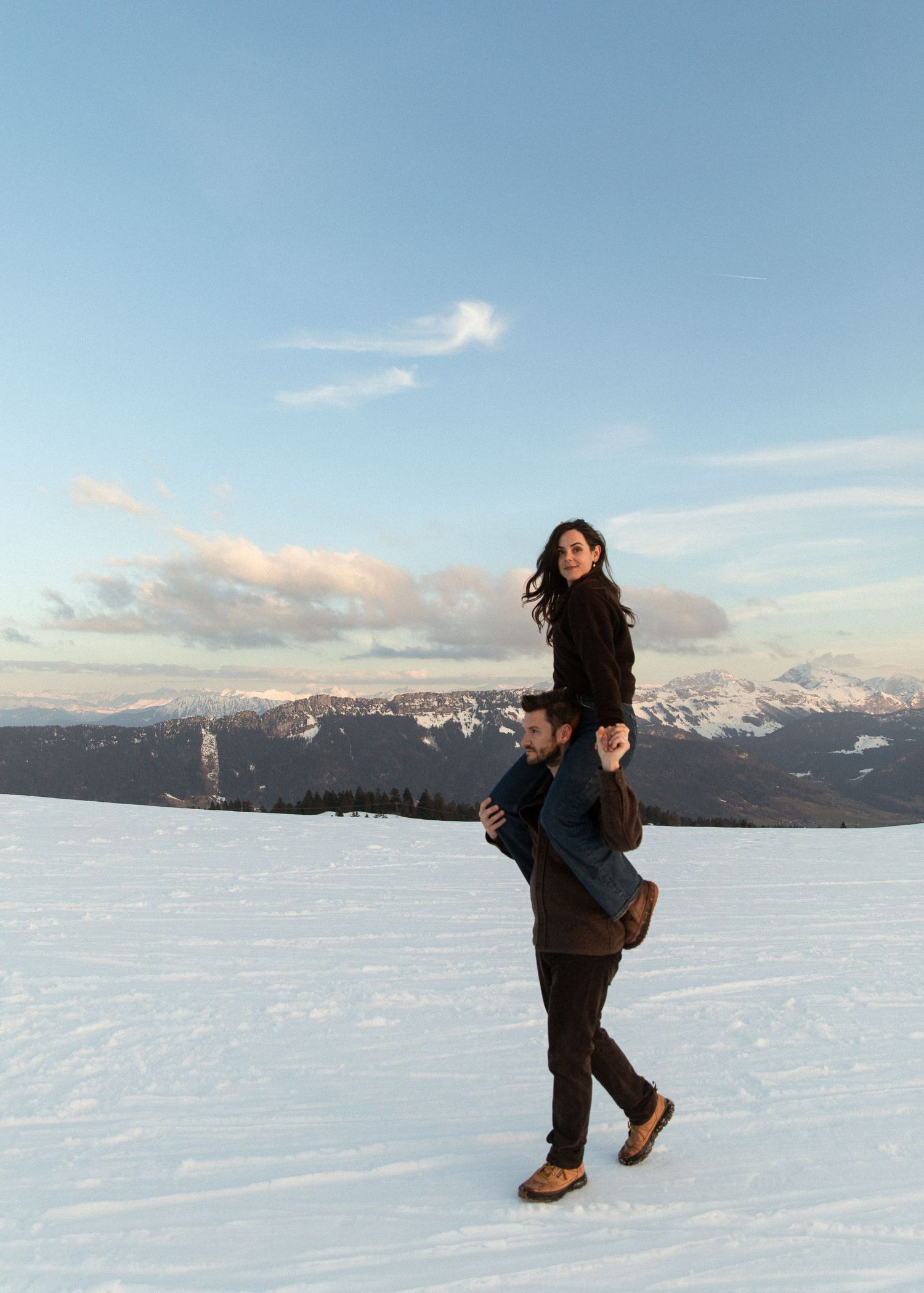 Two people share a playful moment on snowy mountain terrain with snow-capped peaks and blue sky in the background.