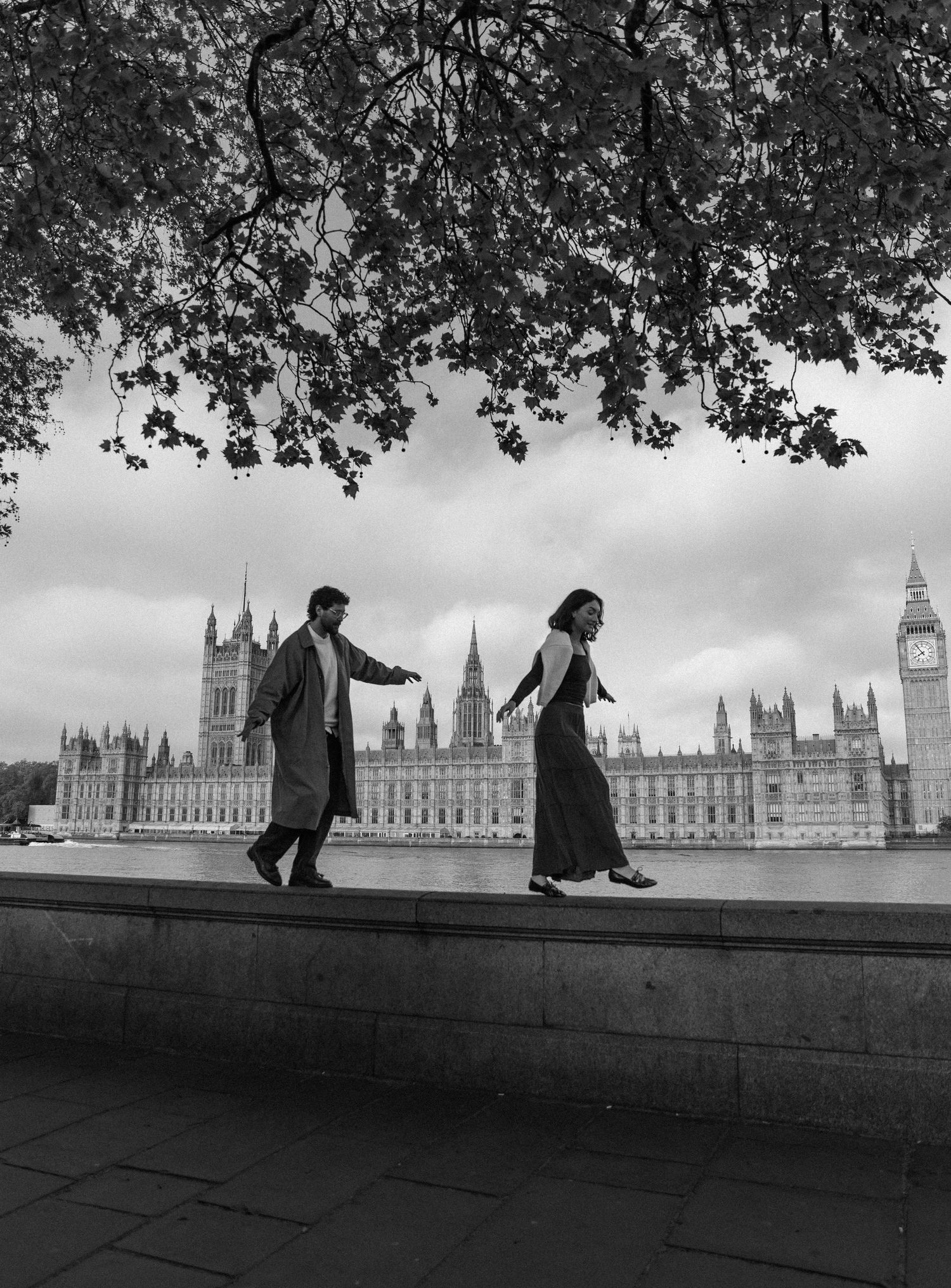 Black and white photo of a couple walking on a wall by the Thames with the Houses of Parliament and Big Ben behind them.