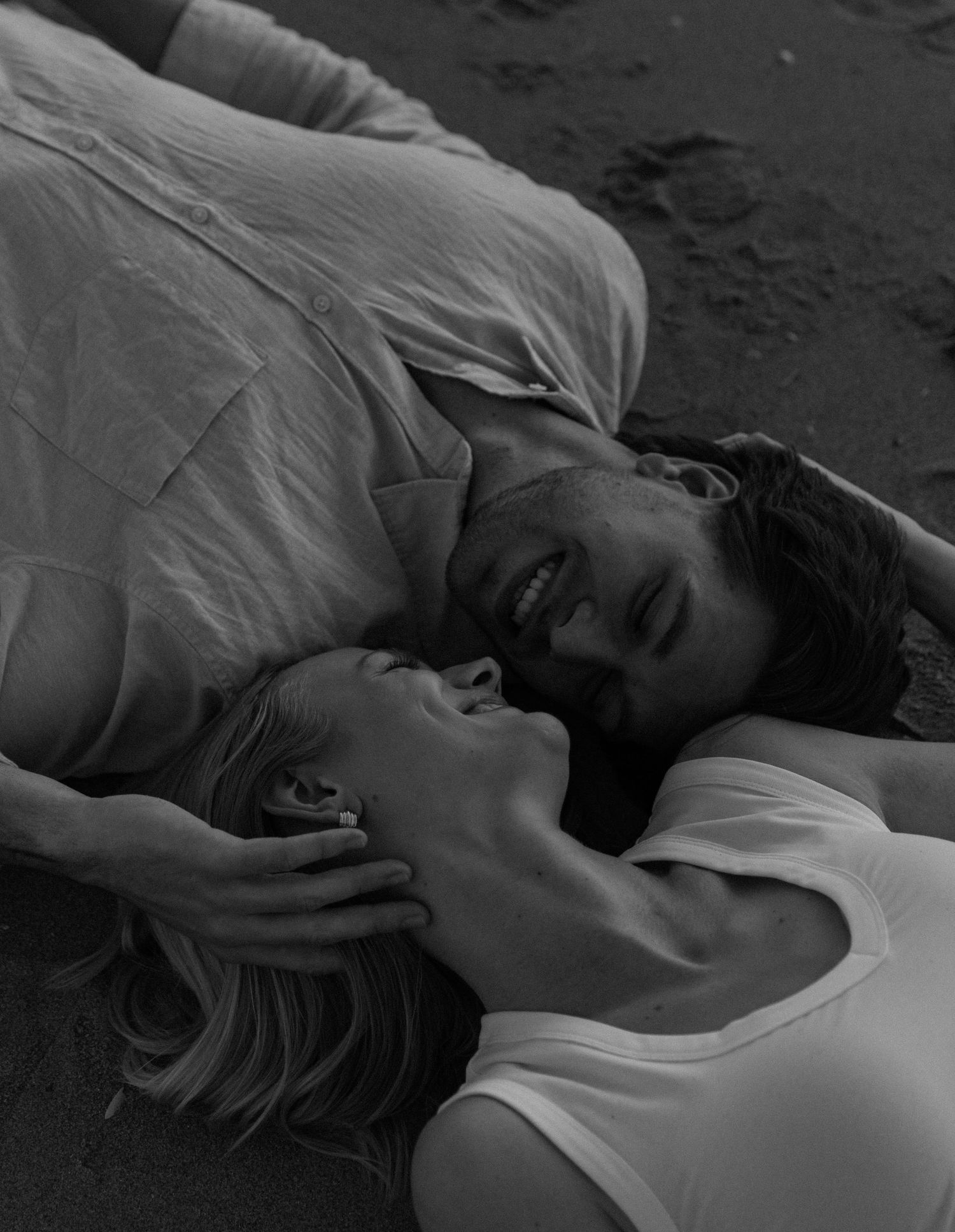 Black and white photo of a smiling couple lying on a beach, gazing at each other romantically.