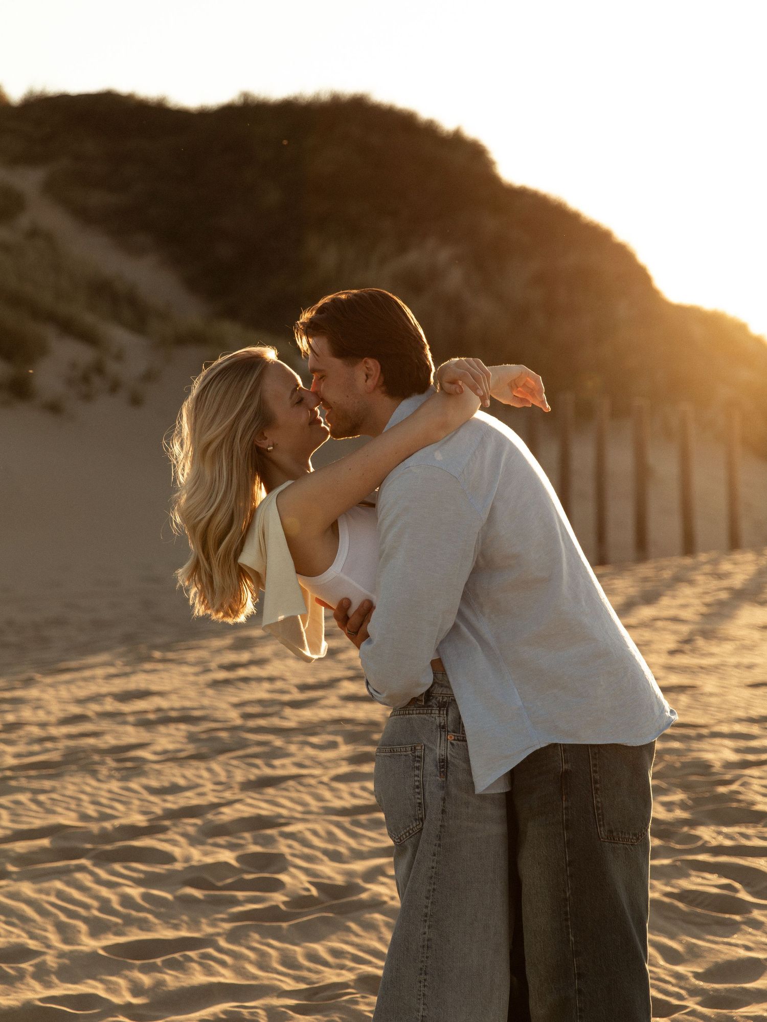 Couple embracing and kissing on a sandy beach at golden sunset with sand dunes in the background.