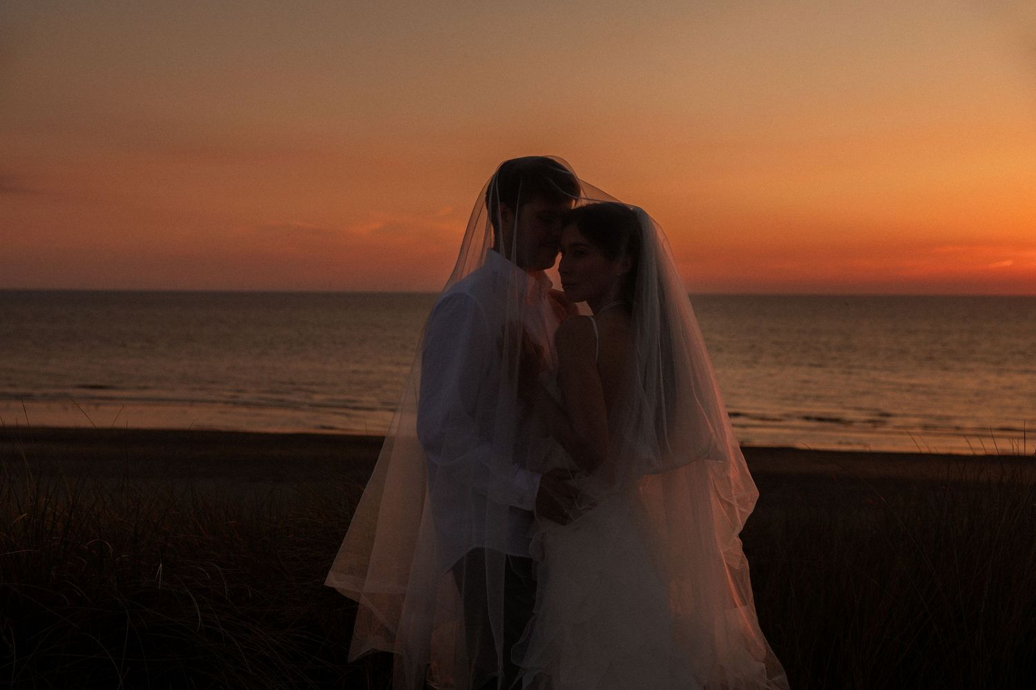 Bride and groom share a romantic moment on the beach at sunset, silhouetted against a vibrant orange sky.