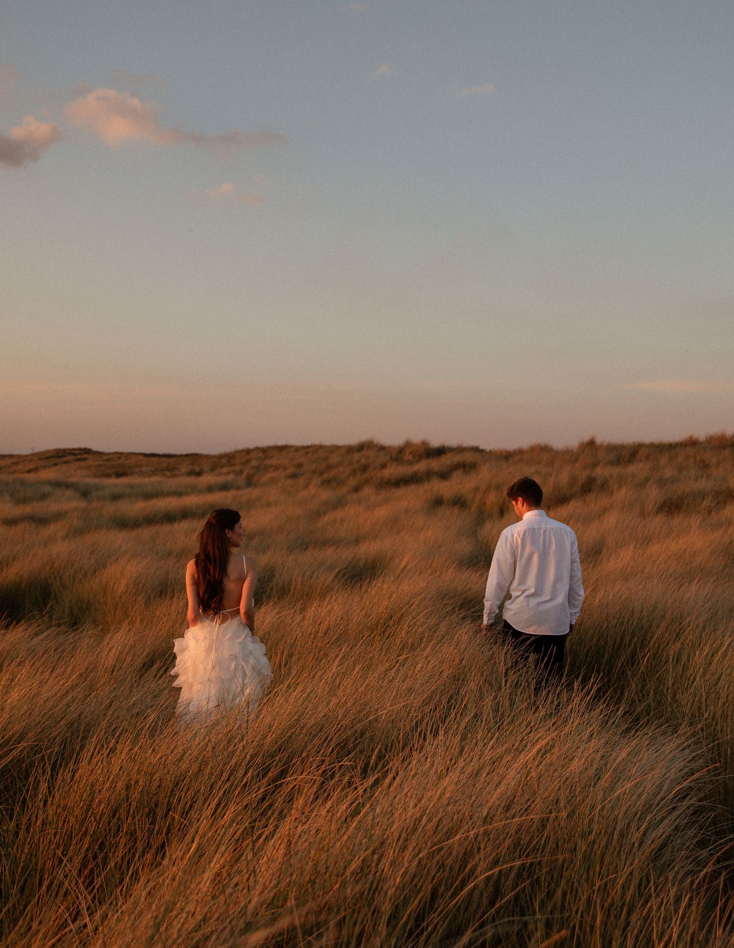 A bride and groom walk through golden grass dunes at sunset, backs to camera, in a romantic outdoor setting.