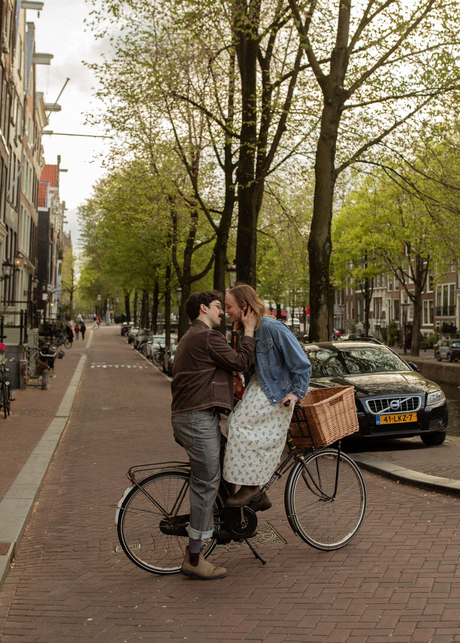 A romantic couple sharing a kiss while standing with a bicycle on a tree-lined Amsterdam street.