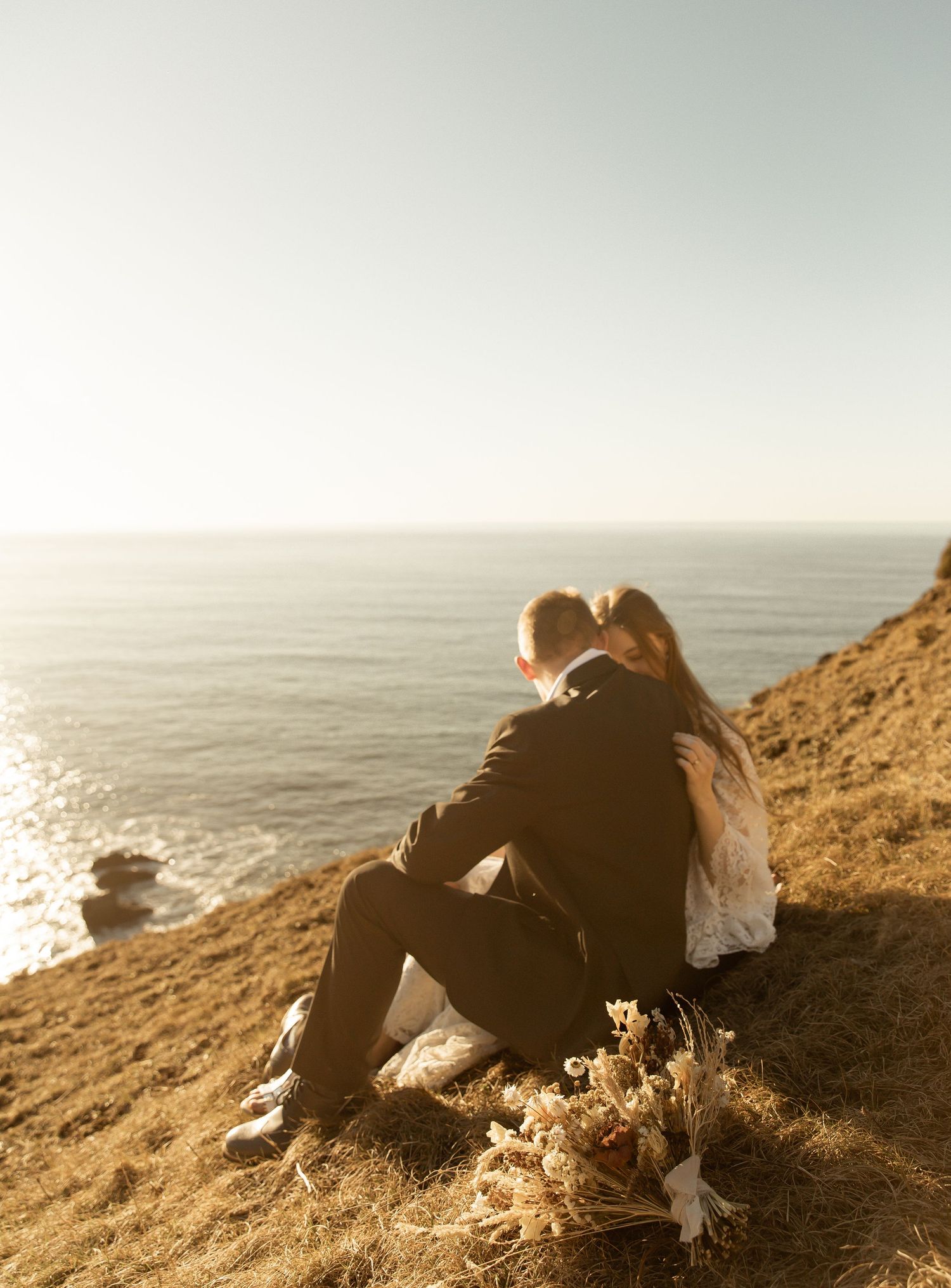 A couple sits together on a rocky coastal cliff overlooking a misty ocean at sunset.