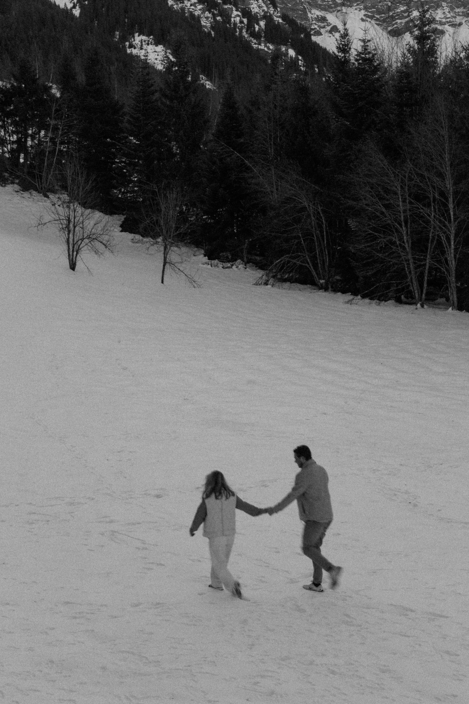 A couple holds hands while walking through a snowy mountain landscape in winter.