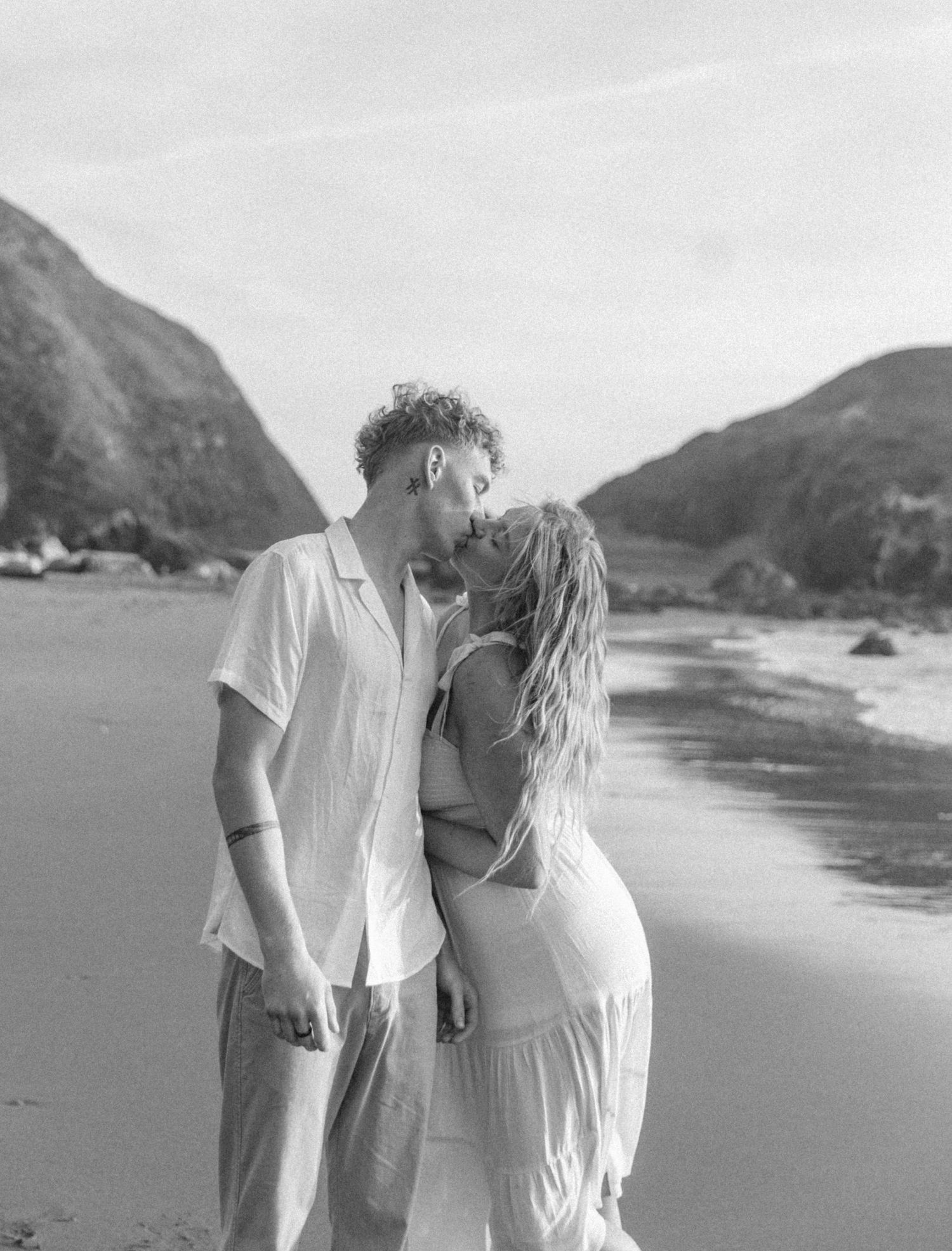 A couple in white outfits shares an intimate moment together while walking along a scenic beach with mountains in the background.