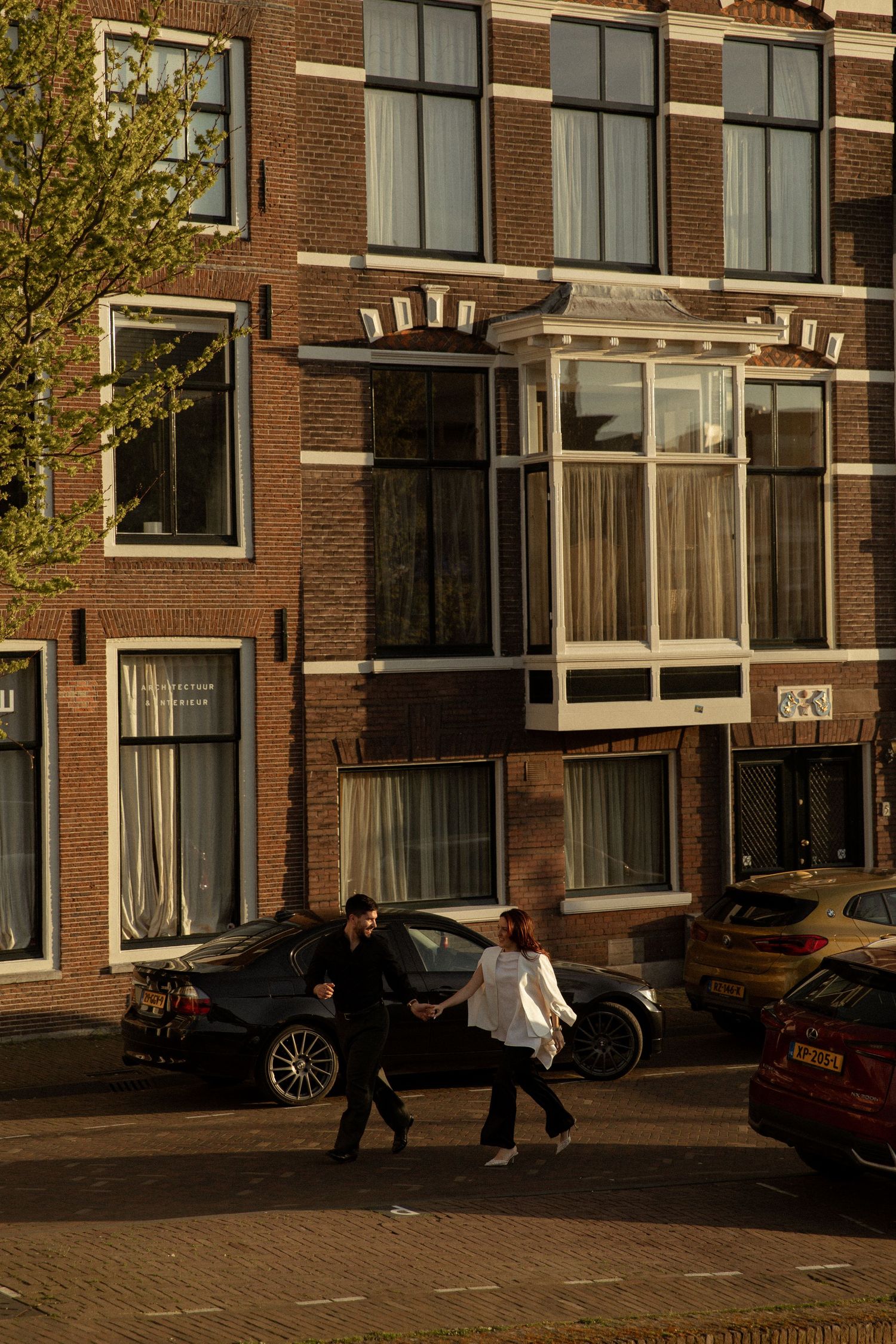 Couple walking hand in hand on a sunny street in Amsterdam, with classic Dutch brick buildings and parked cars in the background.