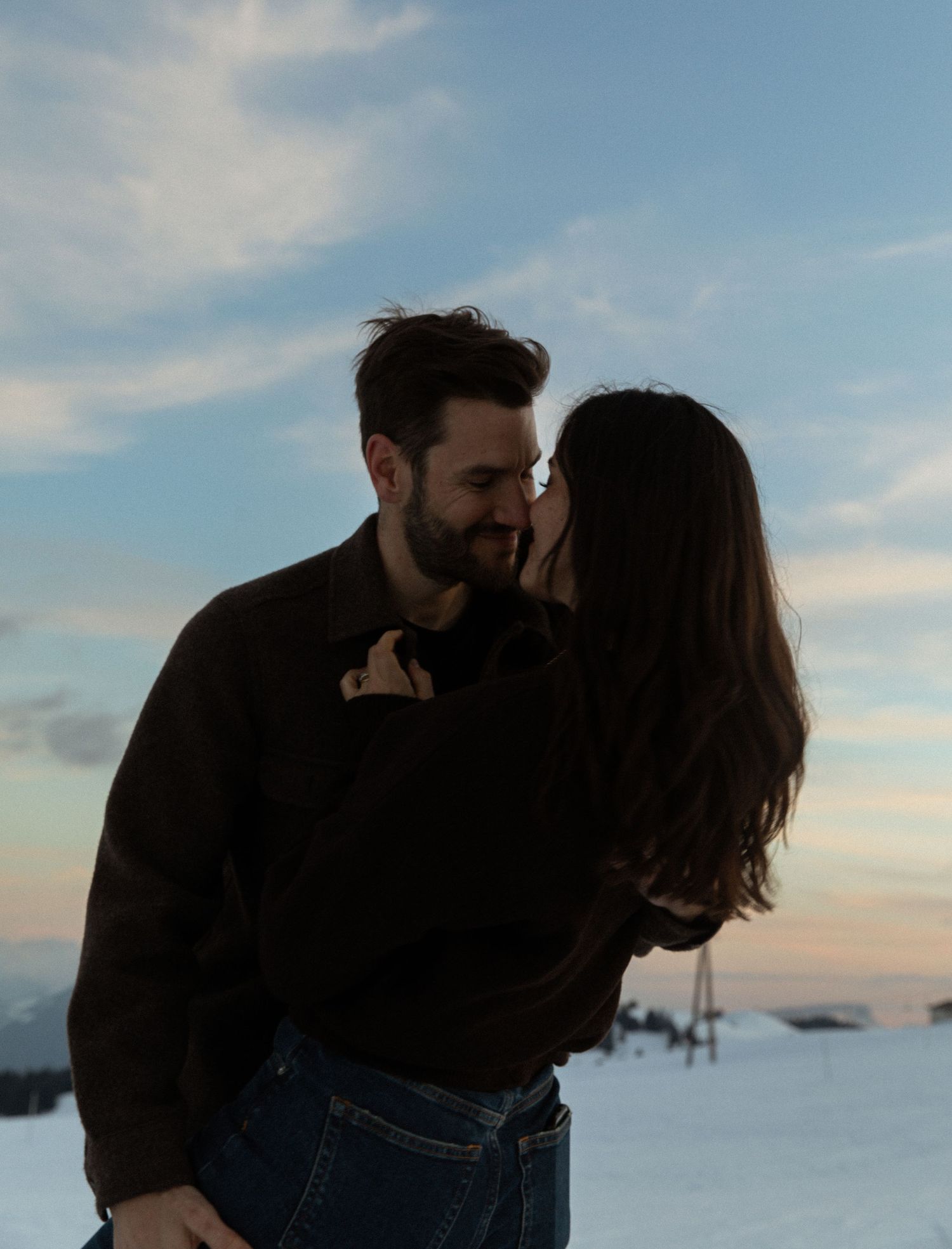 Silhouetted couple embracing during a snowy winter sunset with a cloudy sky backdrop.