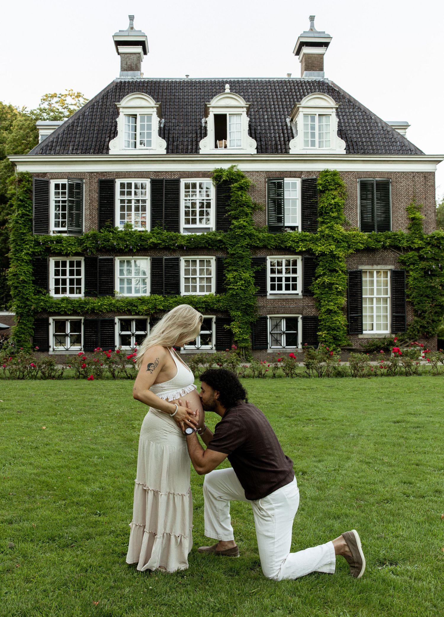 A man kneels kissing a pregnant woman's belly on a lawn before a grand ivy-covered brick mansion.