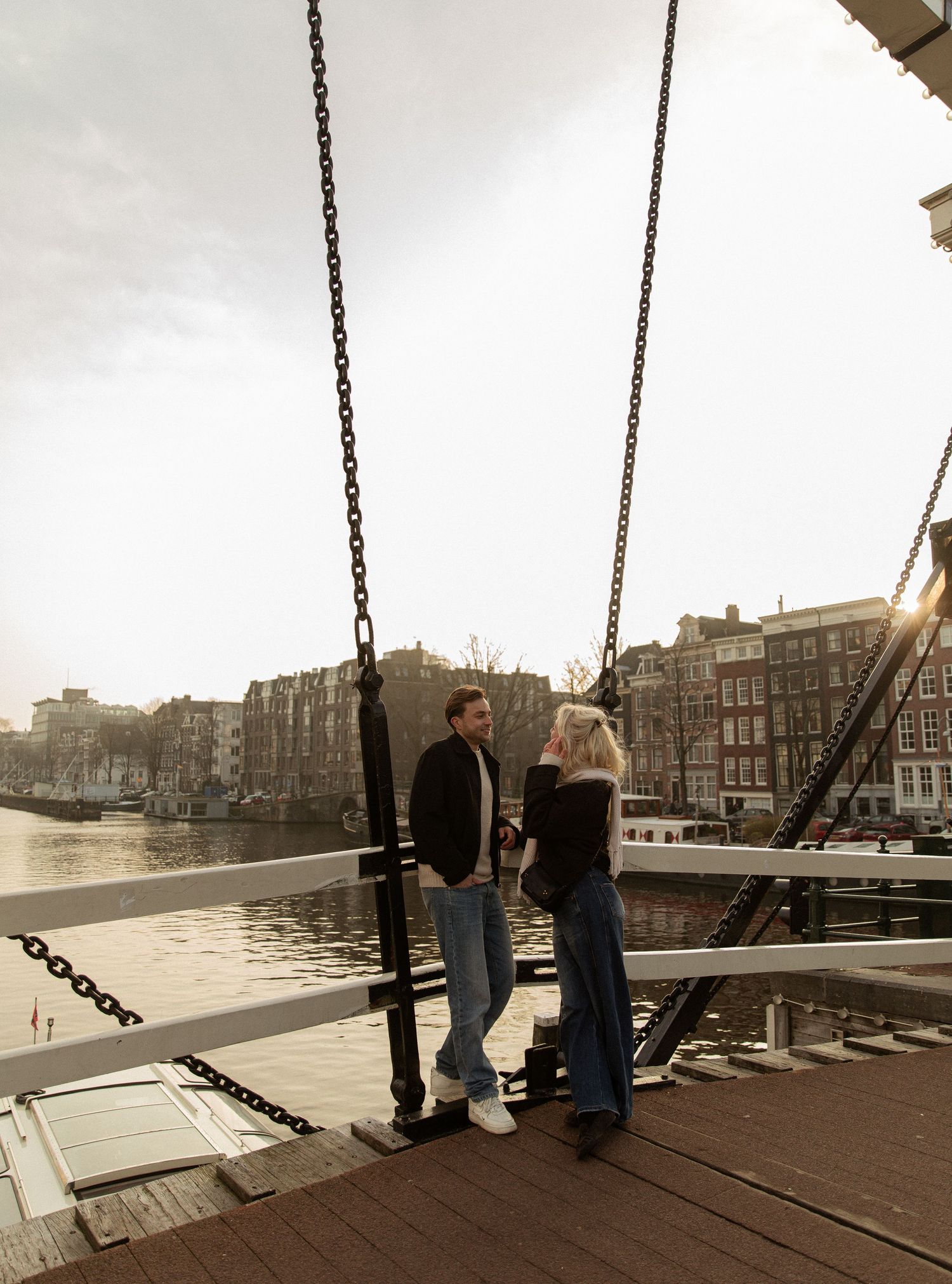 People stand on a wooden deck overlooking a waterfront cityscape with historic buildings and boats at sunset.