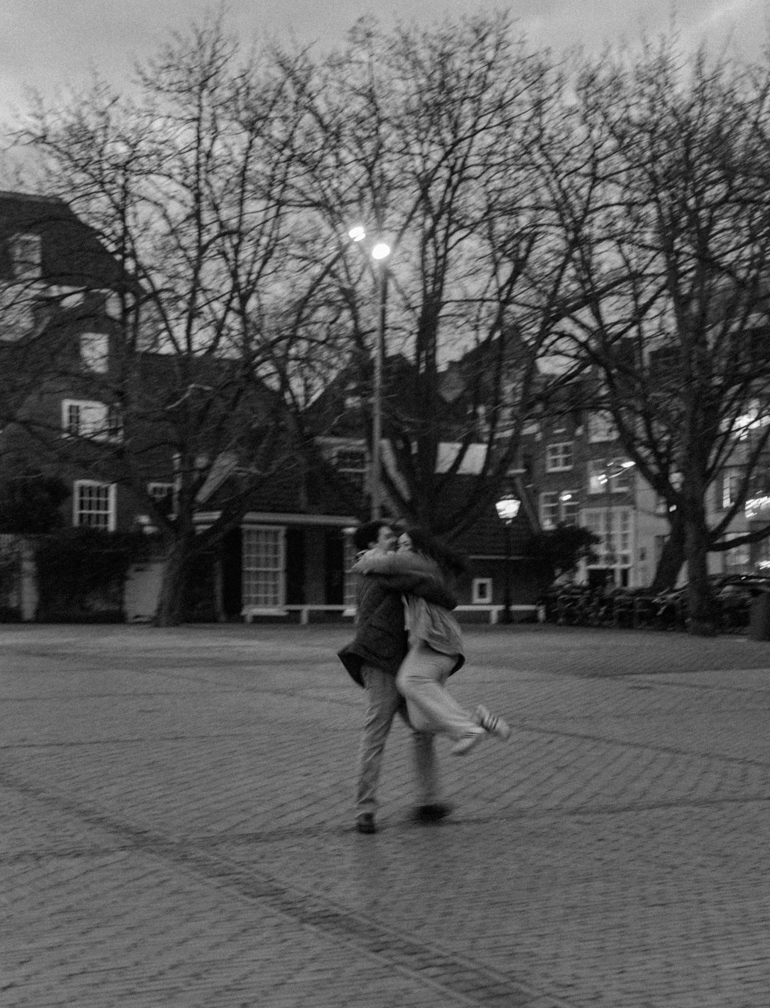 A couple dancing together on a cobblestone plaza at dusk with bare trees and buildings in the background.