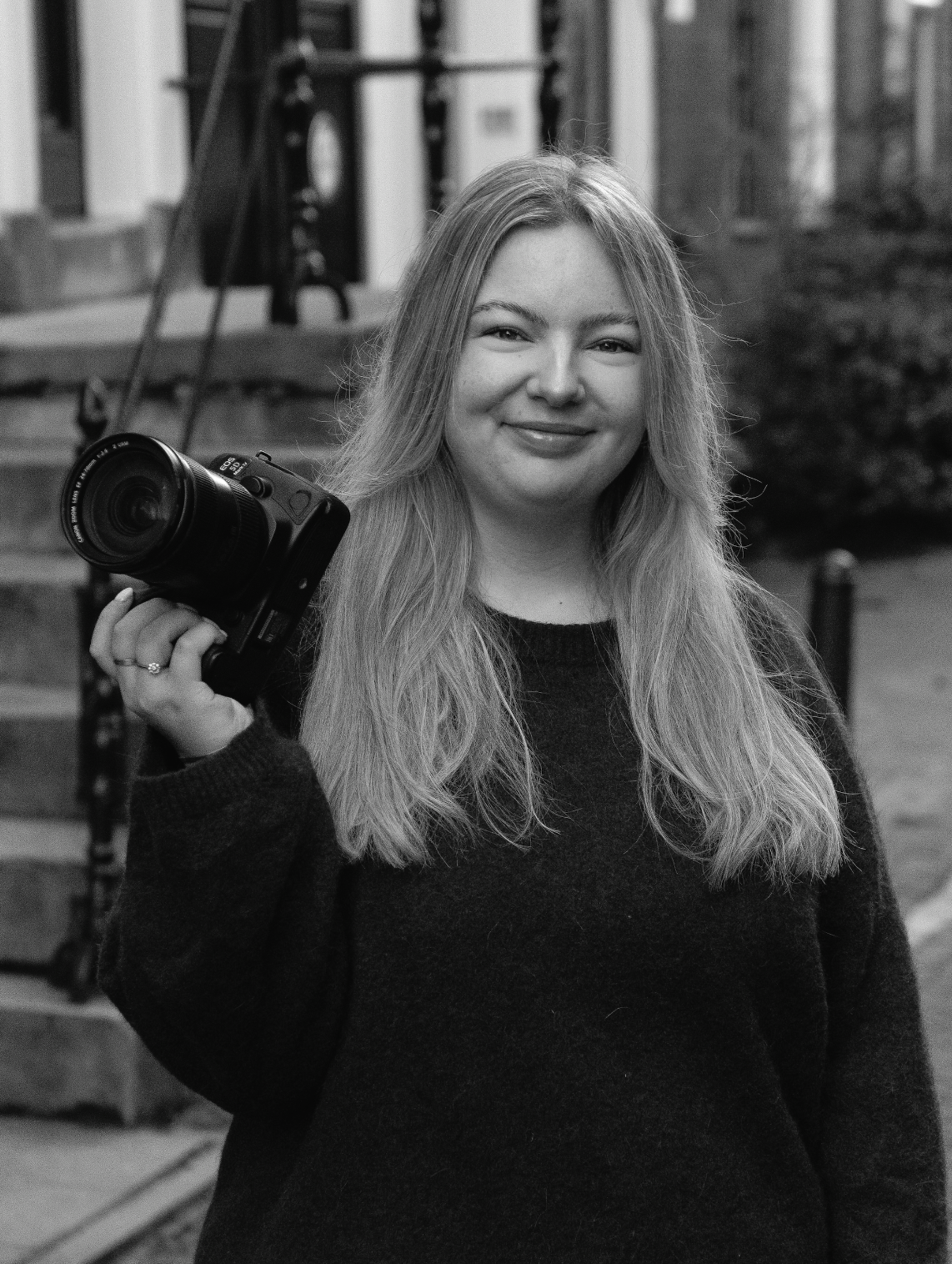 Woman with long blonde hair holding a professional camera, smiling outdoors in a black and white portrait photo.
