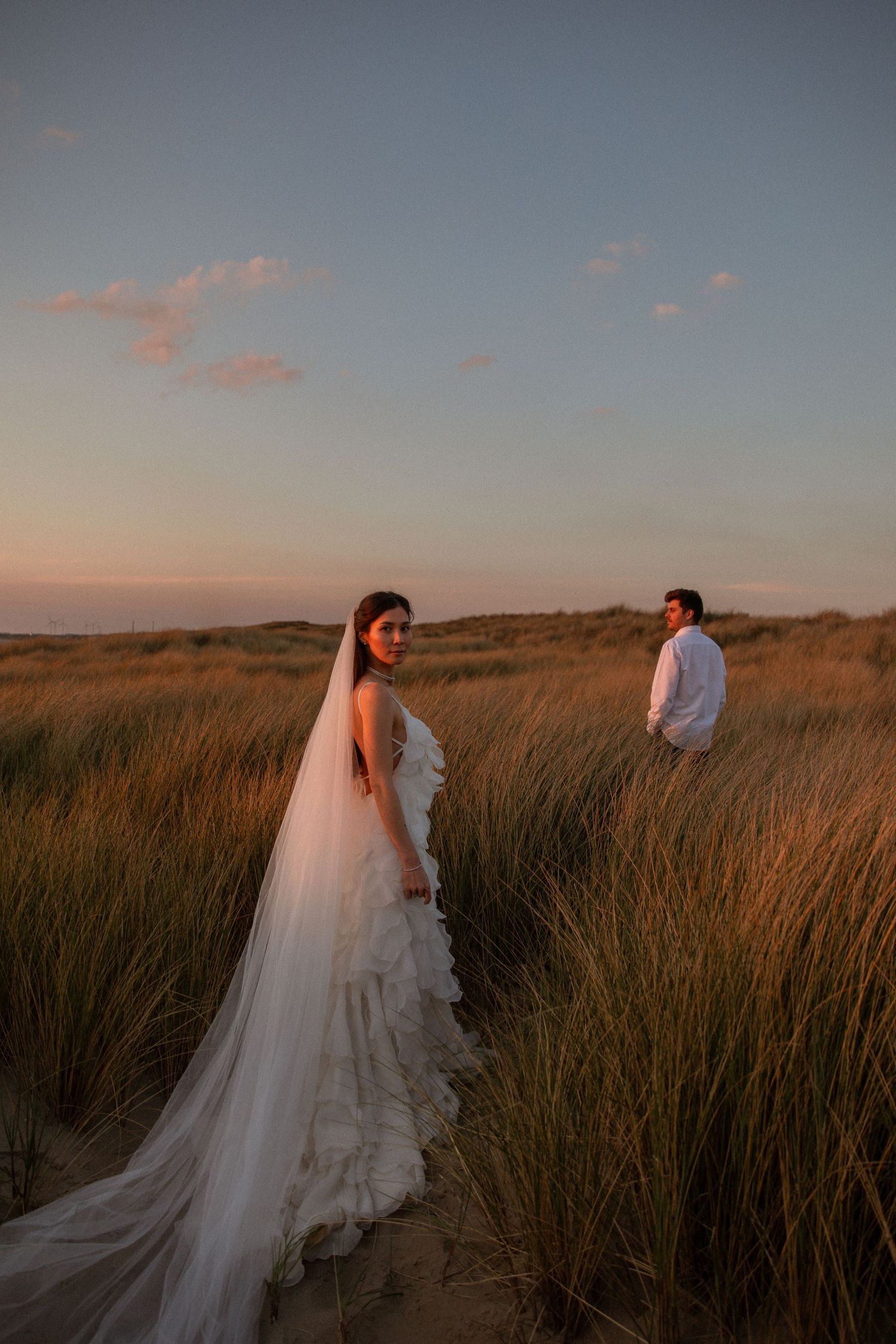 Bride in white ruffled gown and veil stands in golden beach grass at sunset, groom visible in background.