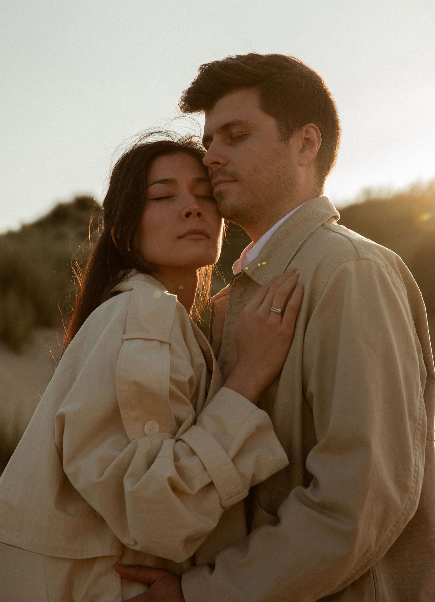 Couple embracing tenderly at golden hour on sandy dunes, wearing matching beige jackets, eyes closed in romantic moment.