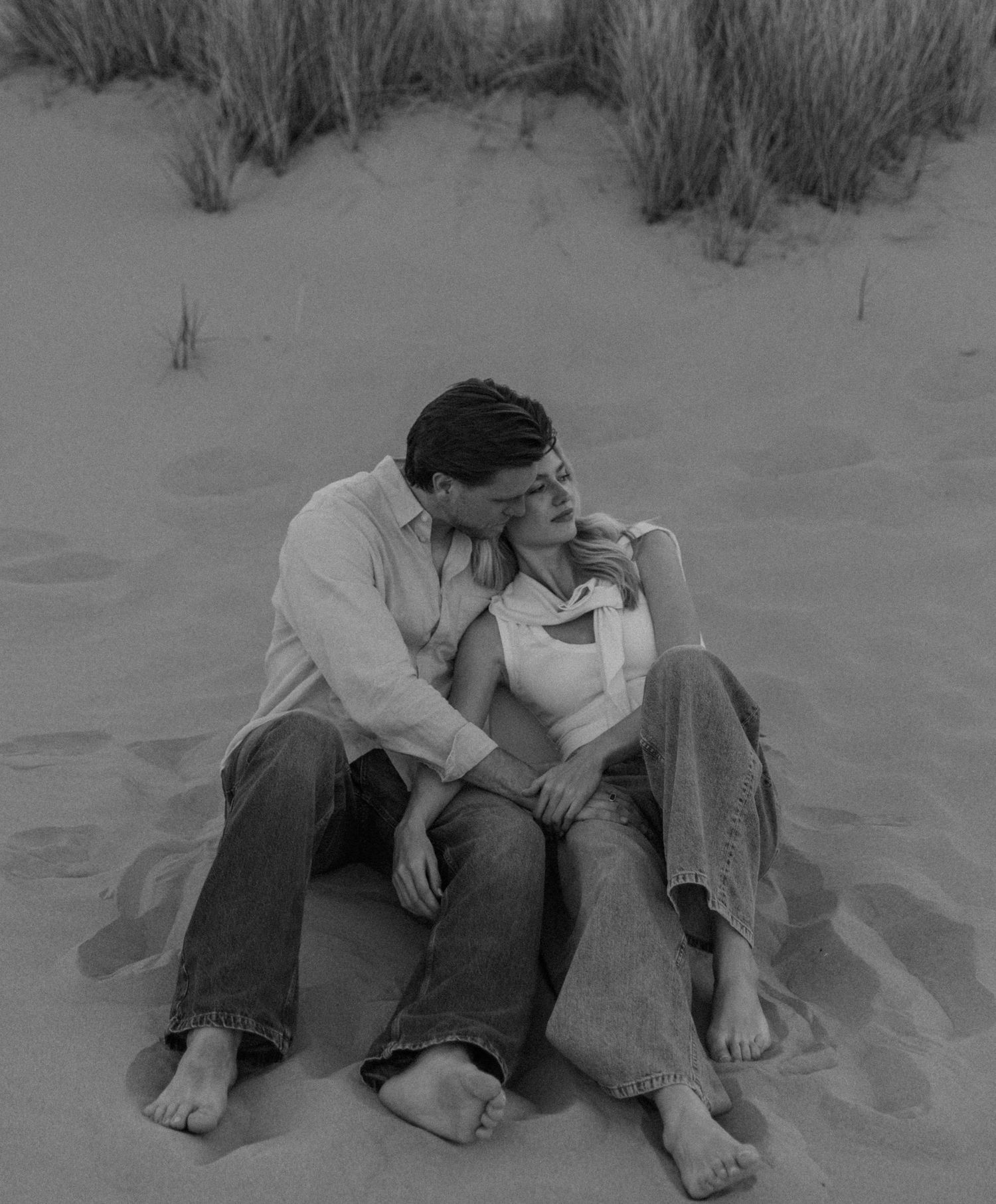 Romantic couple sitting together on sandy beach dunes in black and white photo, sharing an intimate moment.