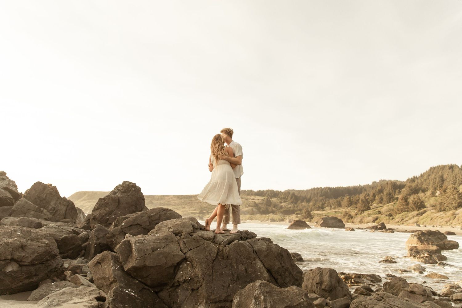 Couple embracing on rocky coastal cliffs at sunset with ocean waves crashing in the background.