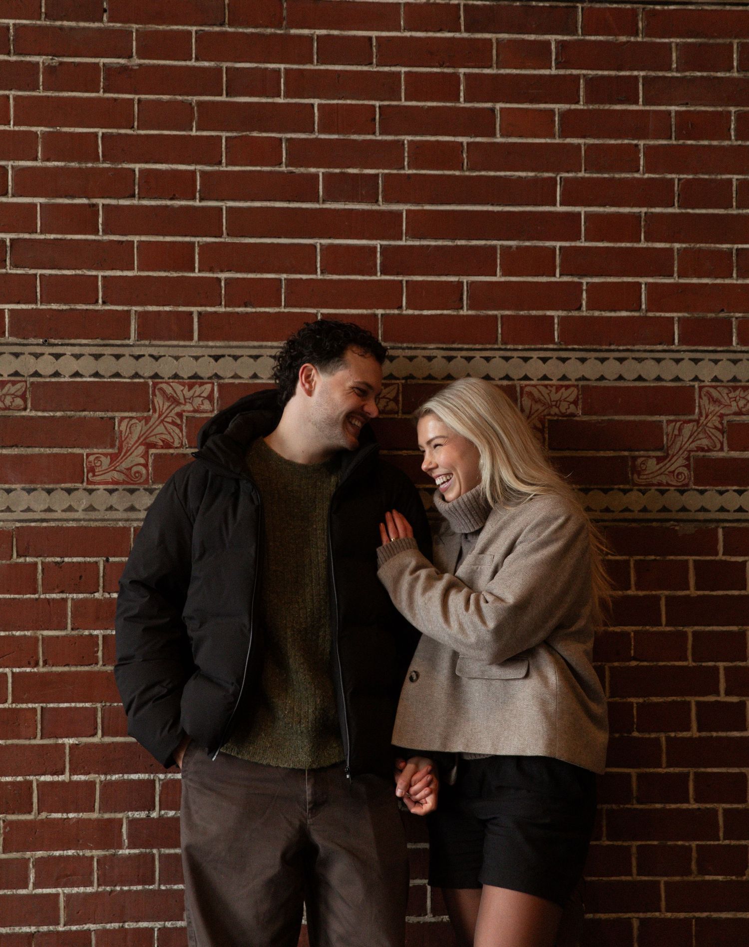 A couple laughs together against a brick wall in a candid photo series showing playful interaction and joy.