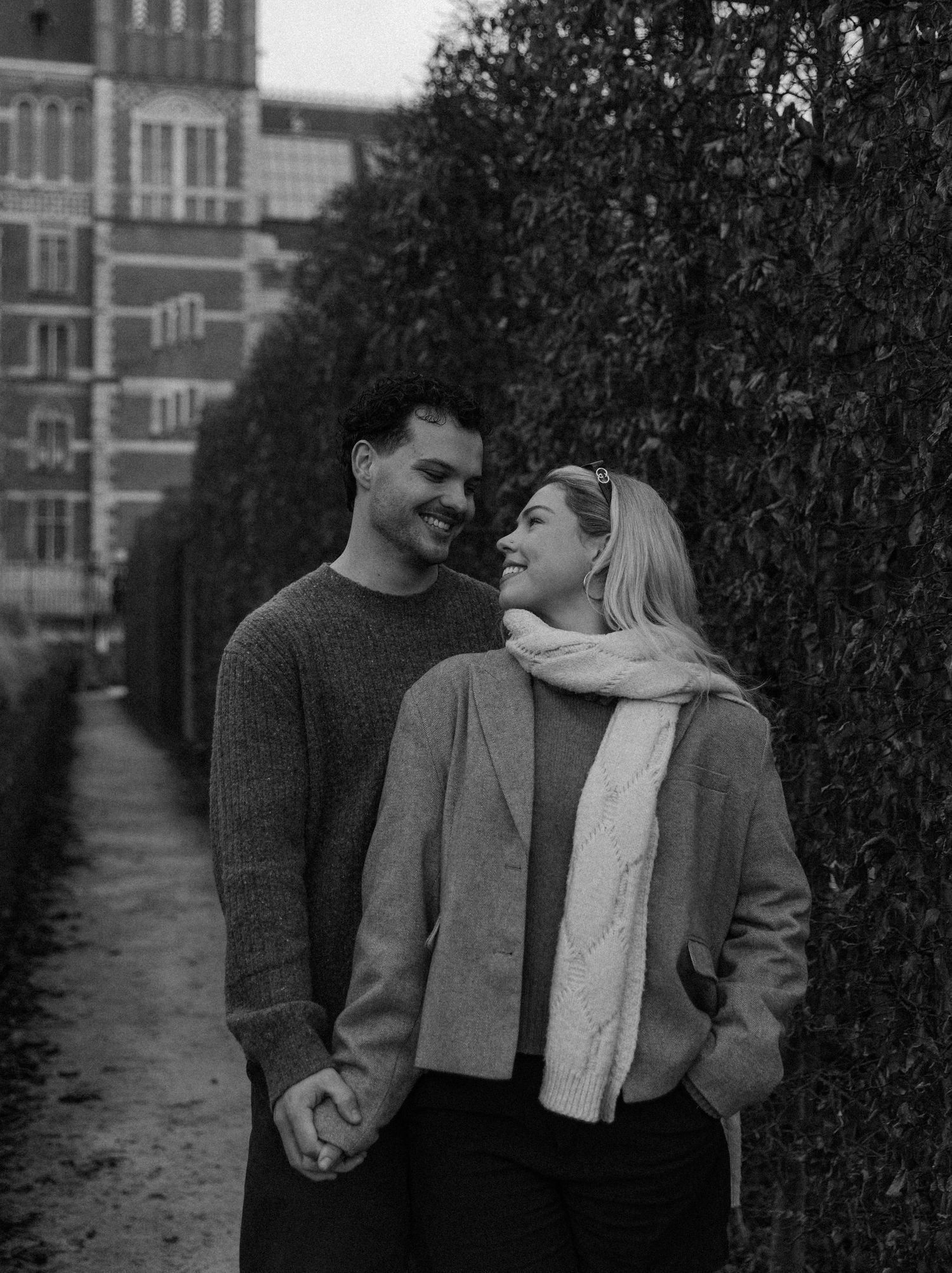 A couple shares a tender moment walking together on a path beside a brick building in black and white.