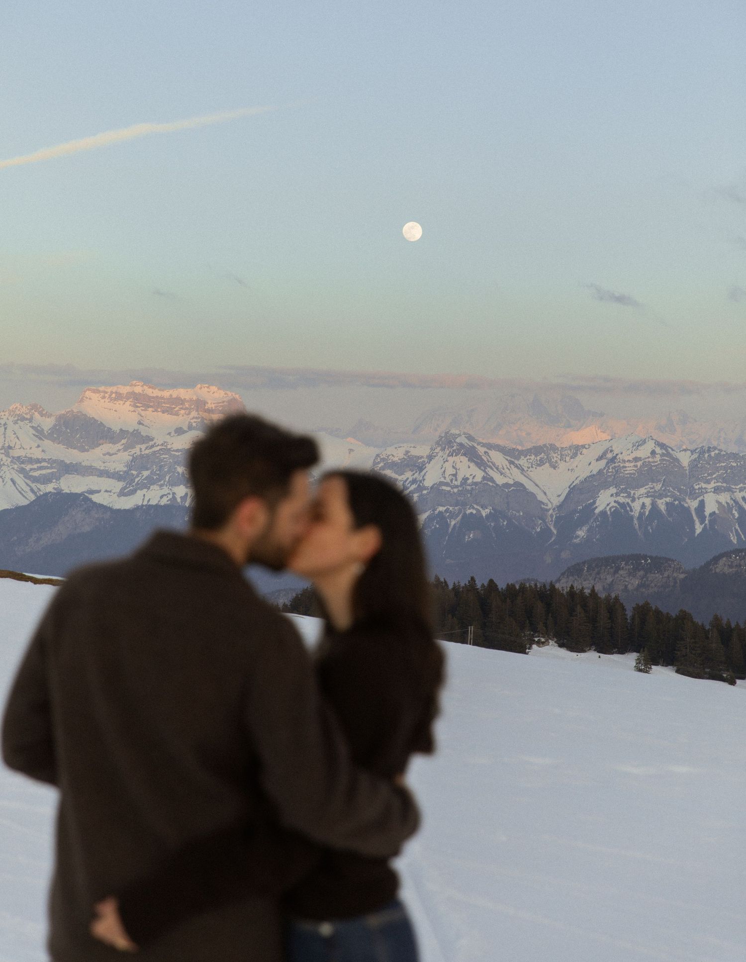 A couple shares a romantic moment against a snowy mountain backdrop with a full moon rising in the winter sky.