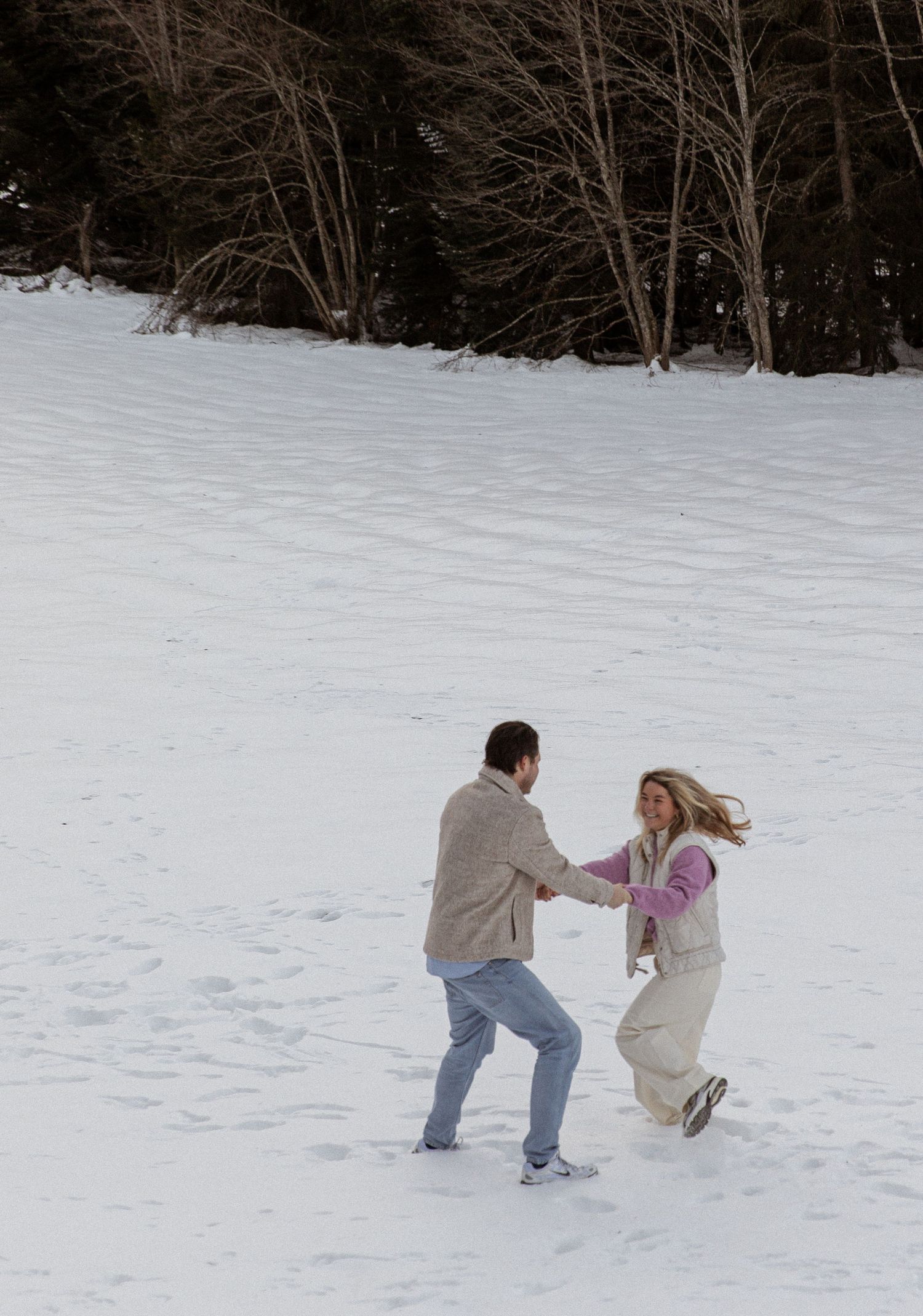 Two people playfully dancing and spinning in the snow with evergreen trees in the background.