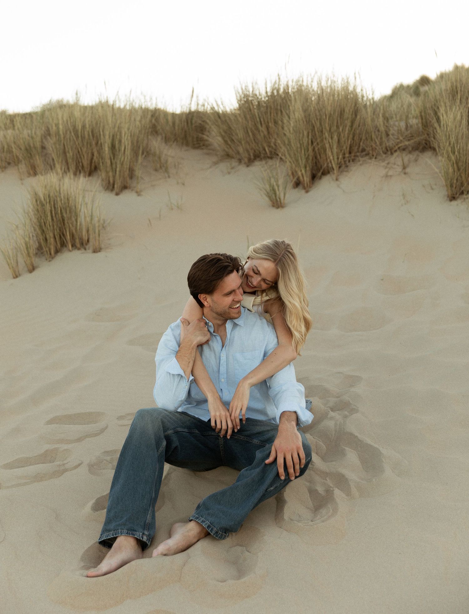 Couple sitting on sandy beach dunes, woman embracing man from behind, both barefoot in casual blue outfits.