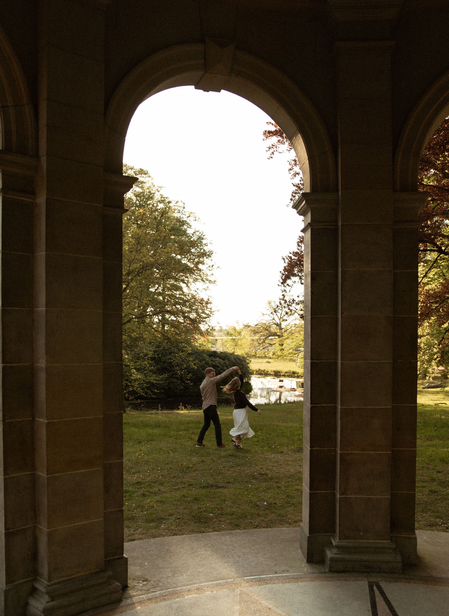 Couple dancing on green lawn viewed through stone archway of historic building with trees and pond in background.