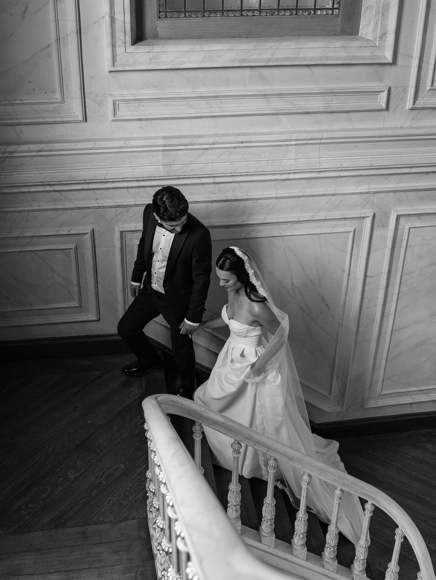 Black and white photo of bride and groom descending elegant marble staircase in grand venue.