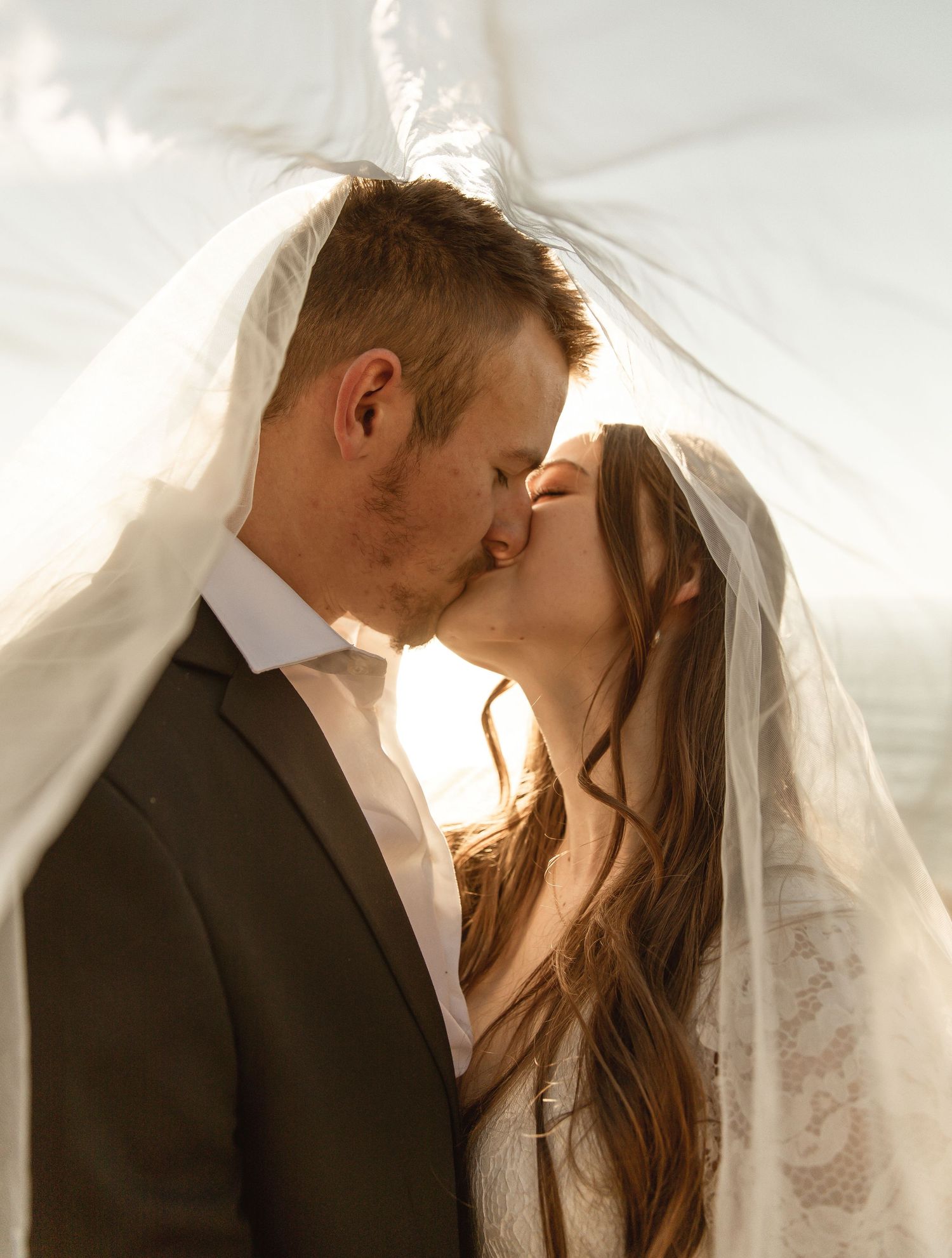 Romantic wedding couple shares intimate moments under flowing white veil in soft natural lighting.