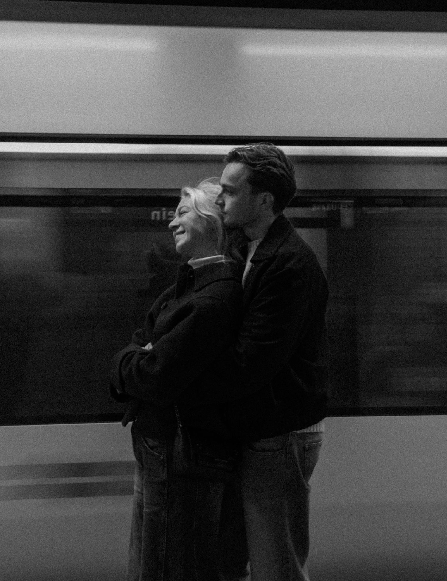A couple embraces on a subway platform in black and white as a train blurs past in the background.
