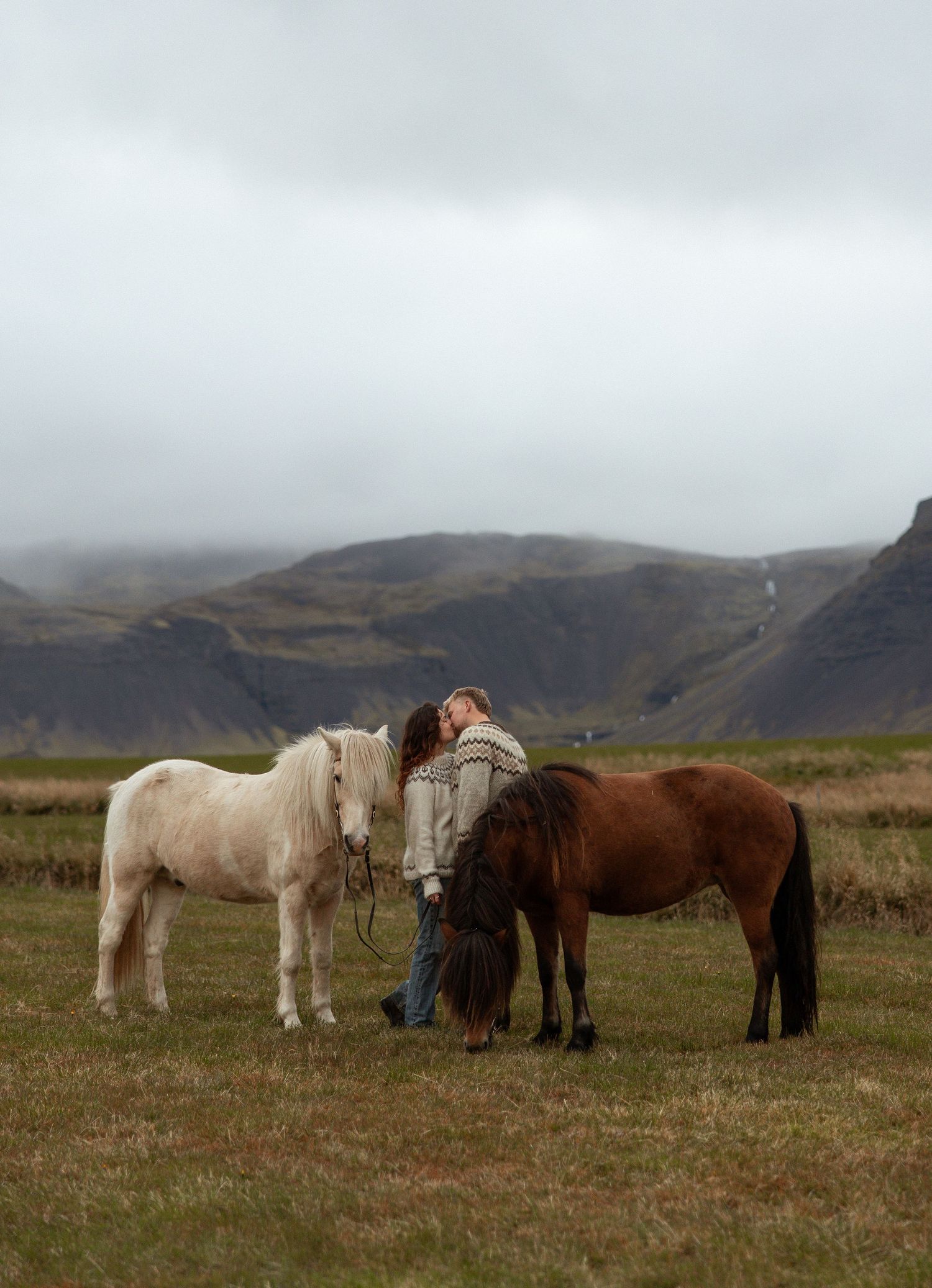 A couple kisses between a white and brown Icelandic horse in a green field with misty mountains in the background.