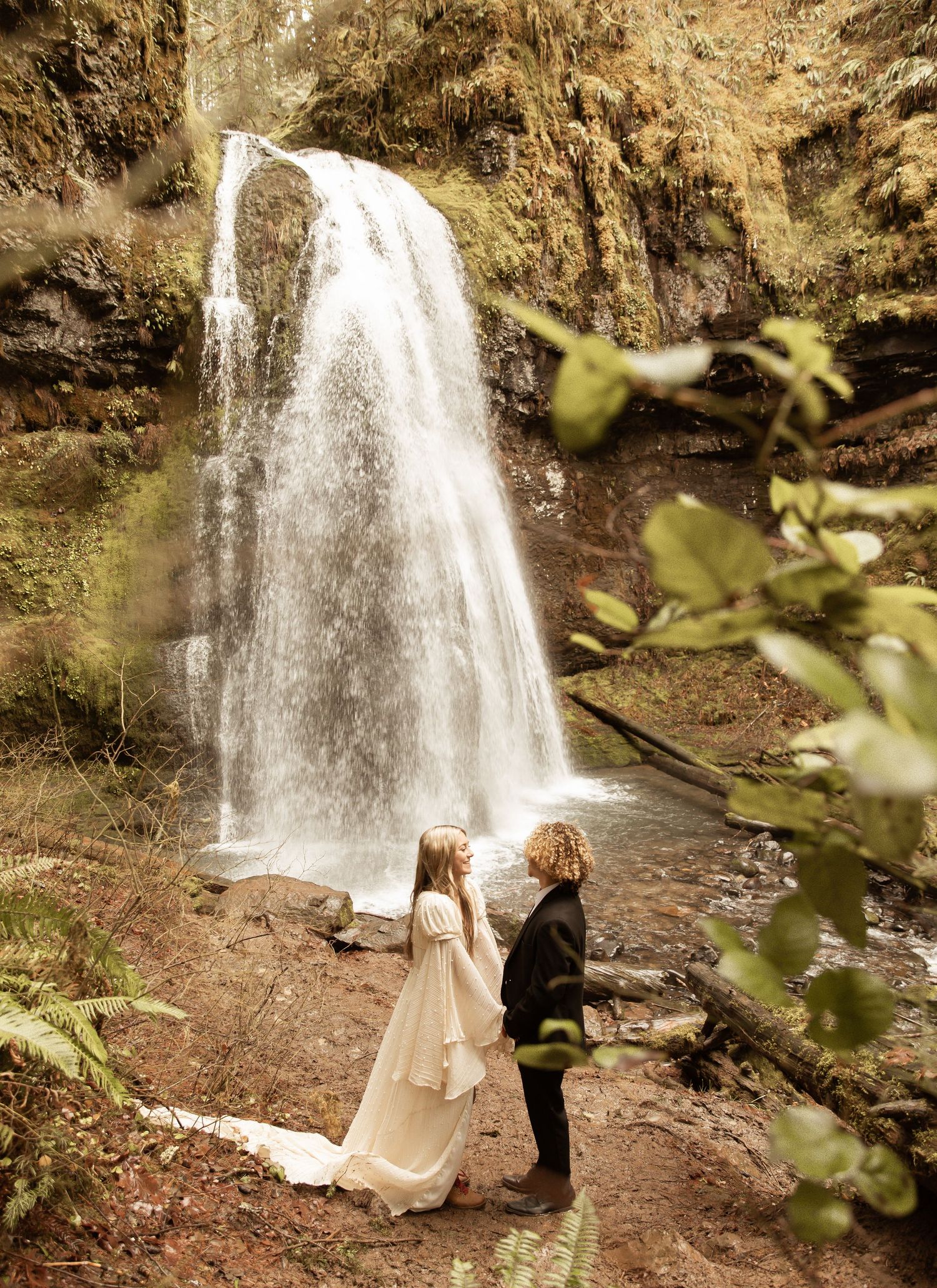 Beautiful waterfall setting frames couple in elegant wedding attire during forest photoshoot.