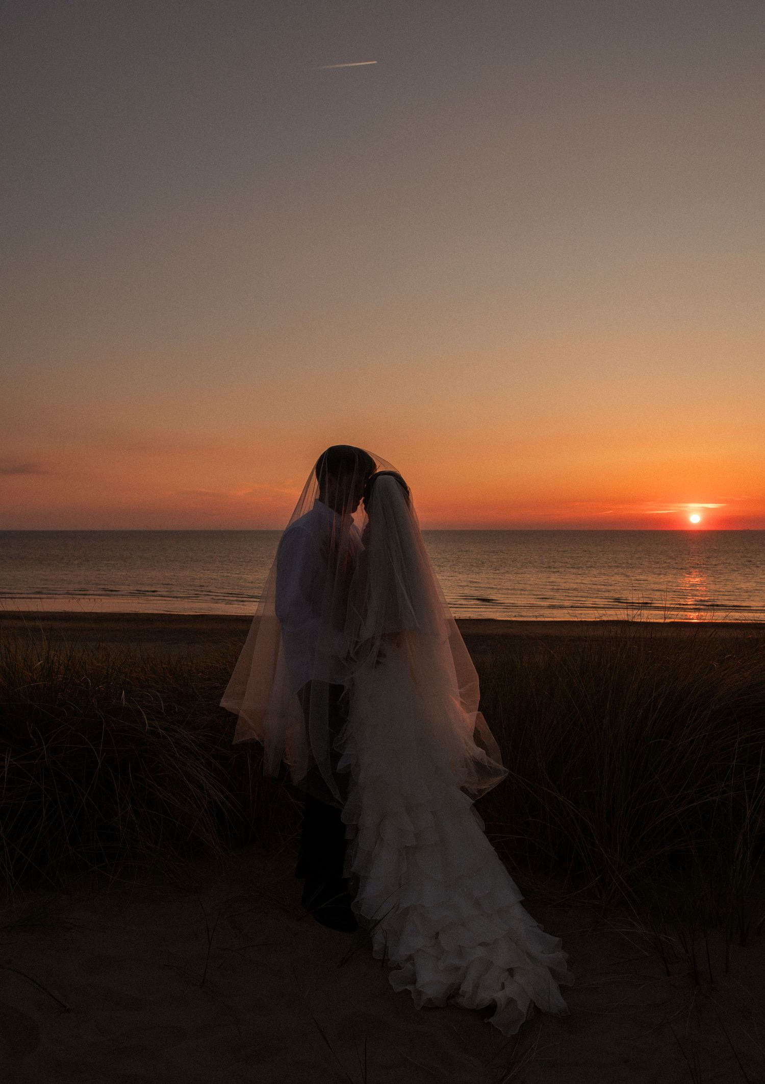 Bride and groom silhouetted against a stunning orange sunset on a beach, bride wearing white gown and veil.