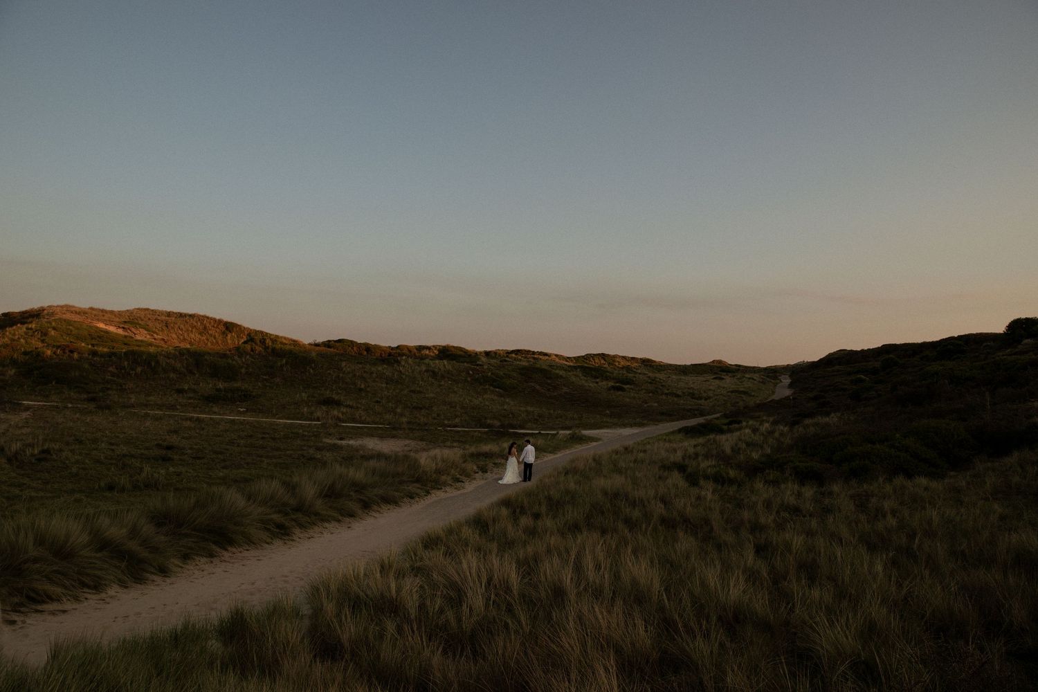 Couple walking on a winding path through green dunes at sunset in a serene landscape.