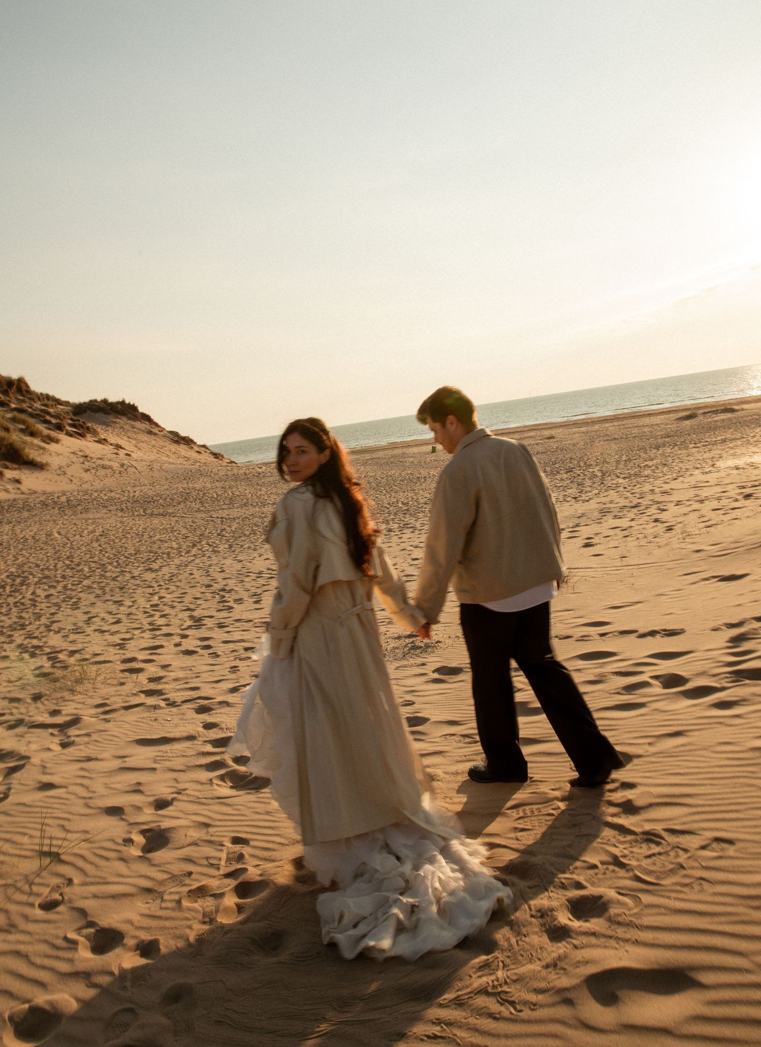 A couple holds hands walking on a sandy beach at sunset, woman in flowing beige dress looks back.