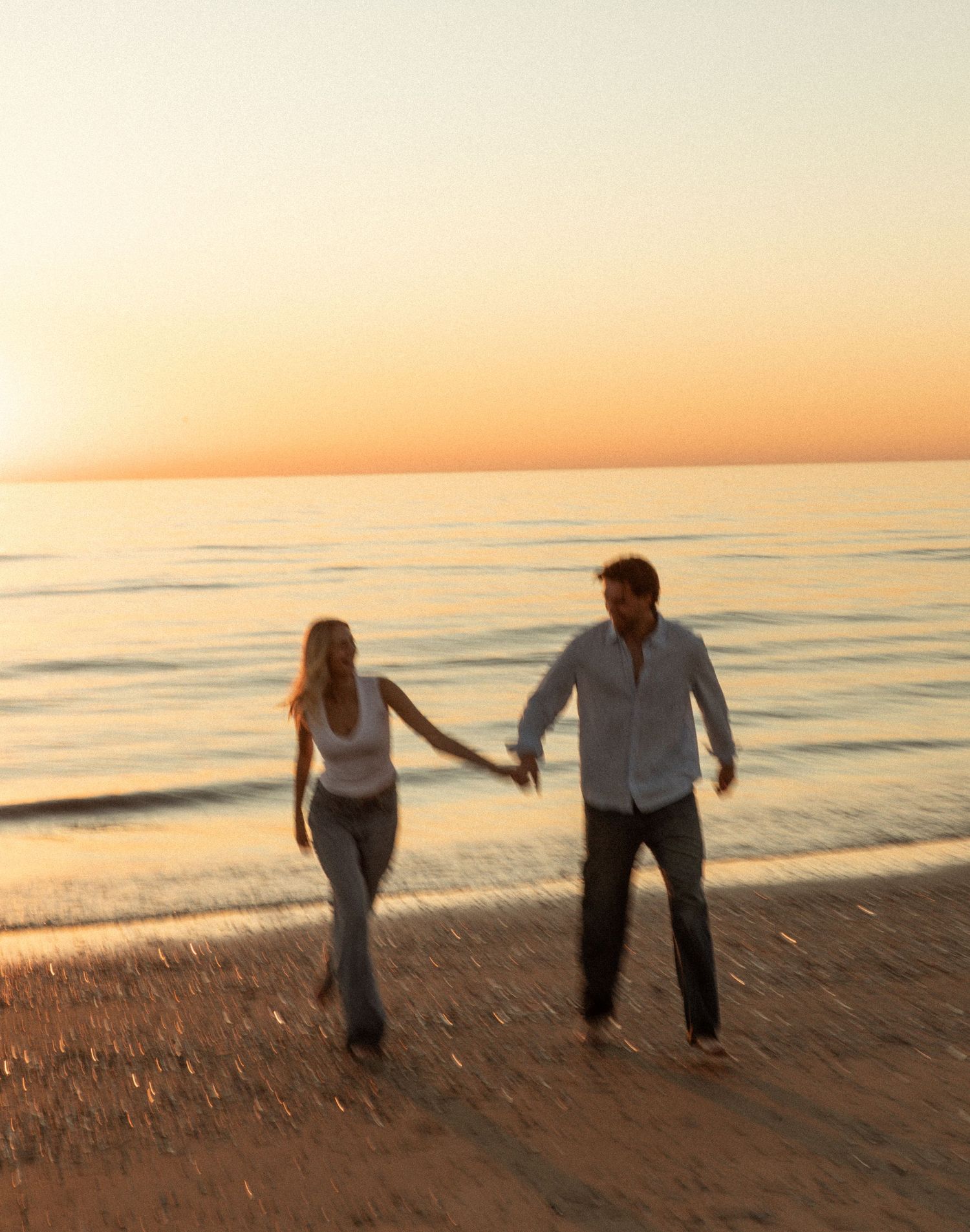 Couple holding hands walking on a sandy beach at sunset with calm ocean waves in the background.