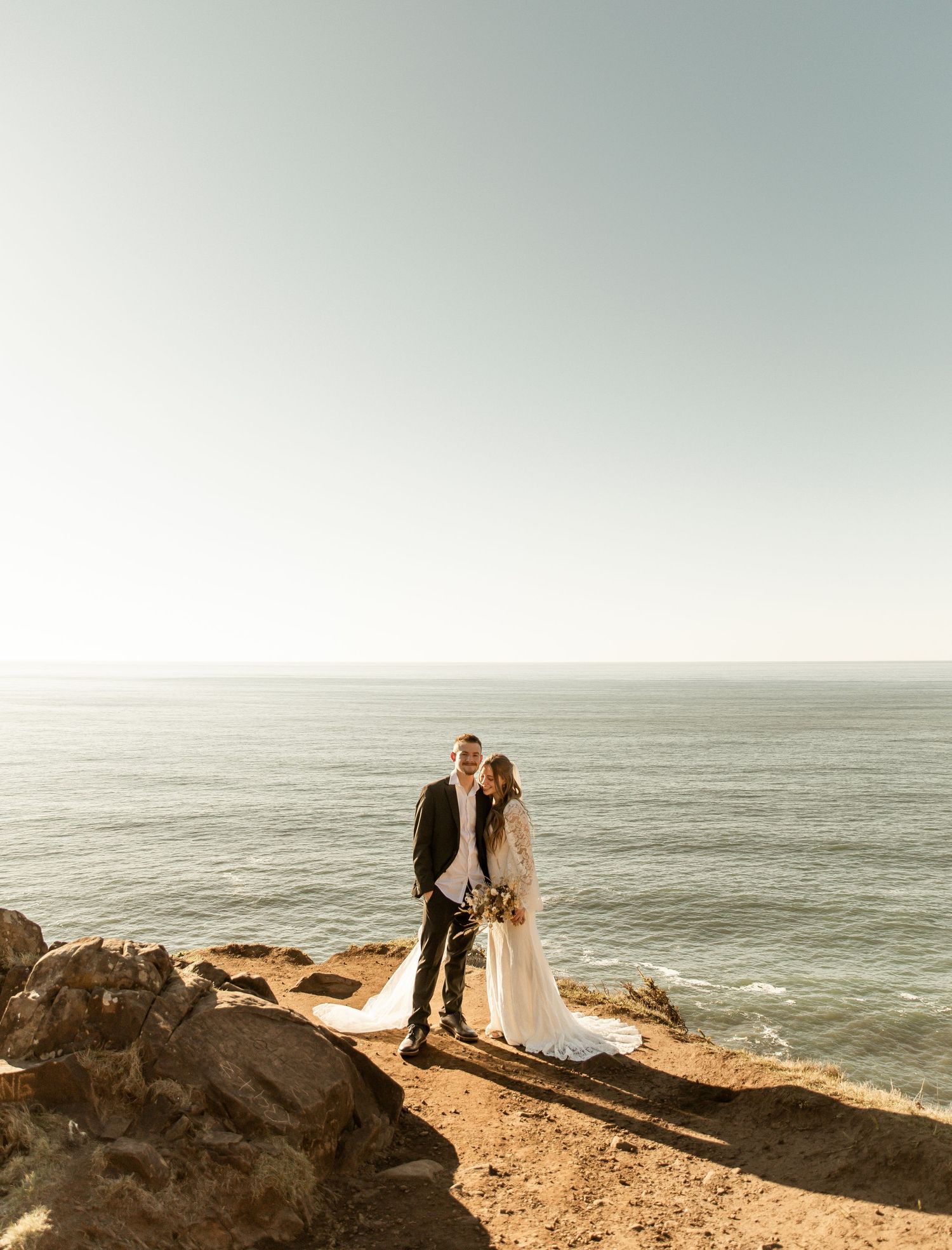 A couple stands together on a dramatic coastal cliff overlooking the Pacific Ocean at sunset.