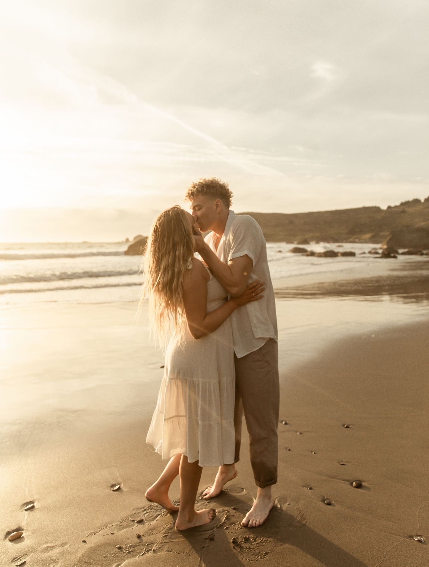 Romantic couple embraces on a sandy beach at sunset with ocean waves and rocky coastline in the background.