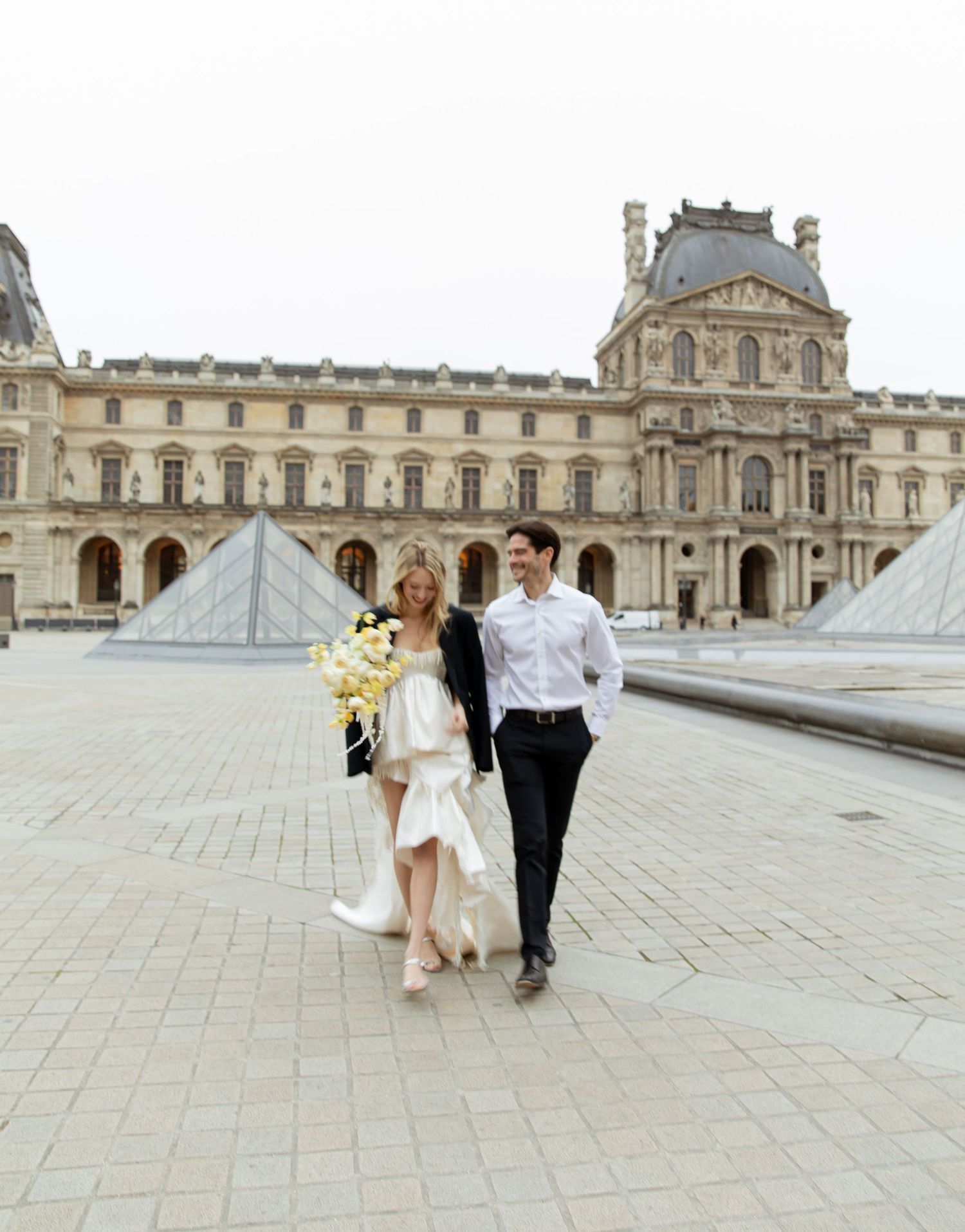 A couple in wedding attire walks near the Louvre pyramid in Paris, bride holding yellow flowers.