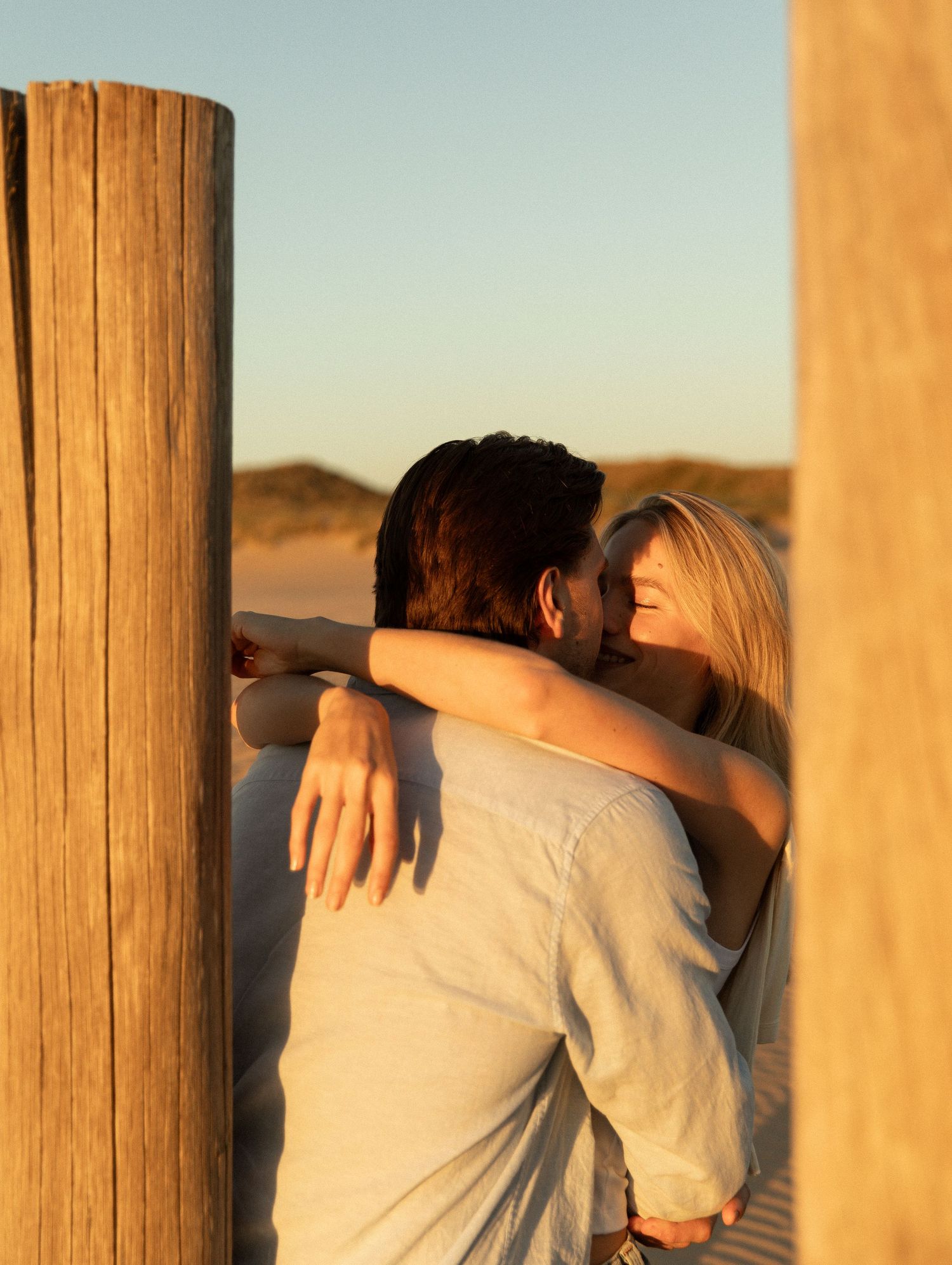 Couple embracing and kissing at golden hour on a sandy beach between wooden posts.
